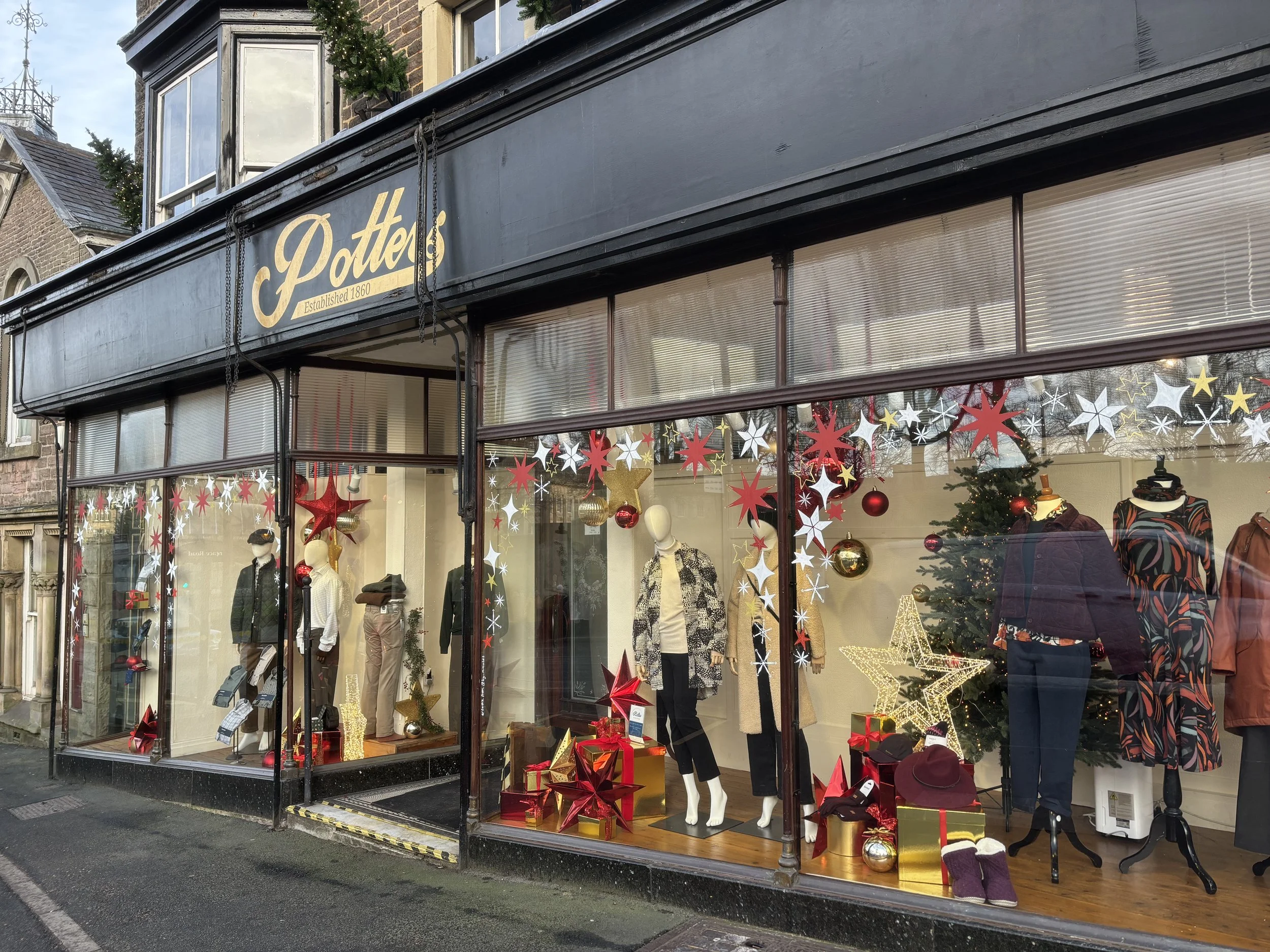 Storefront window display decorated with Christmas ornaments, including stars, baubles, and a Christmas tree, with mannequins dressed in clothing inside. The store's sign reads 'Potters' and was established in 1860.