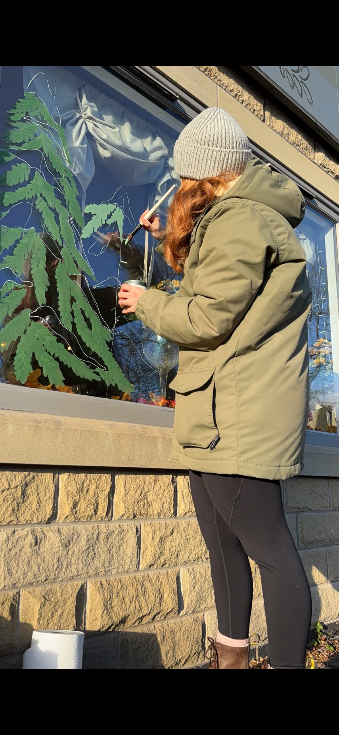 Person with red hair, wearing a gray beanie and tan jacket, painting a mural on a window with a paintbrush. The mural depicts large green leaves.