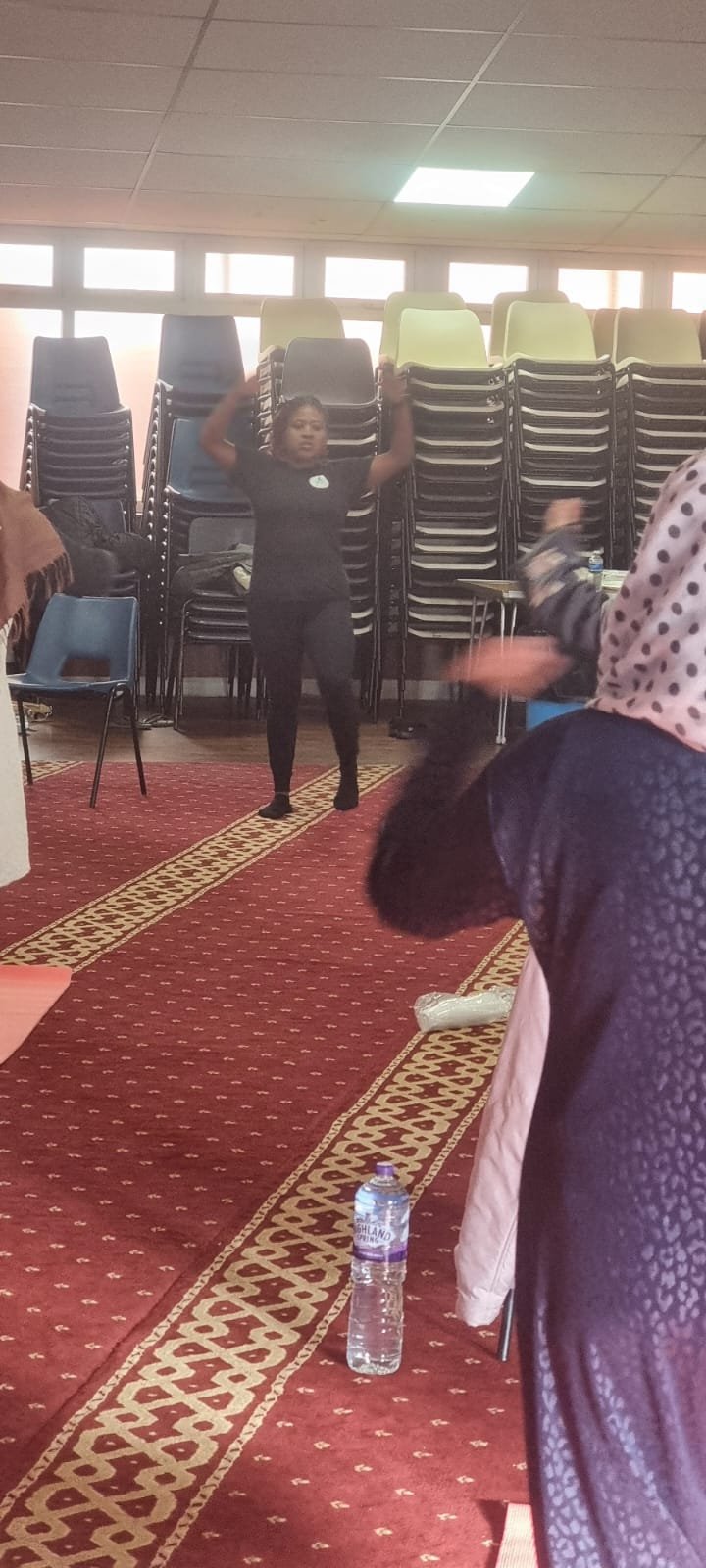 A woman in black pants and a black shirt is standing in front of a stack of chairs, raising her arms. Several people are present, with one partially visible on the right side. The room has a red patterned carpet, plastic water bottle on the floor, an