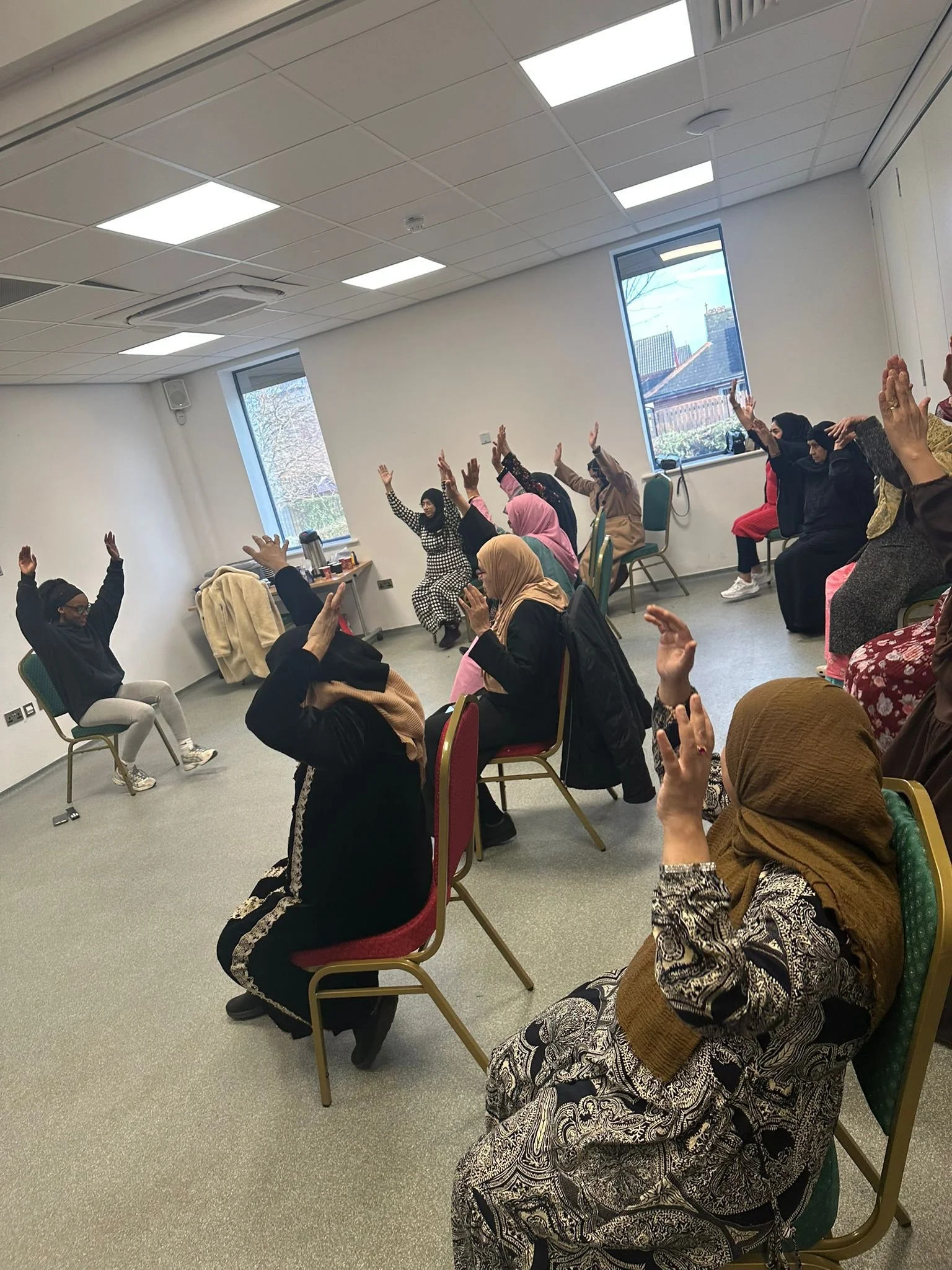 A group of women seated in a room with white walls and large windows, raising their hands in a group activity or meeting.