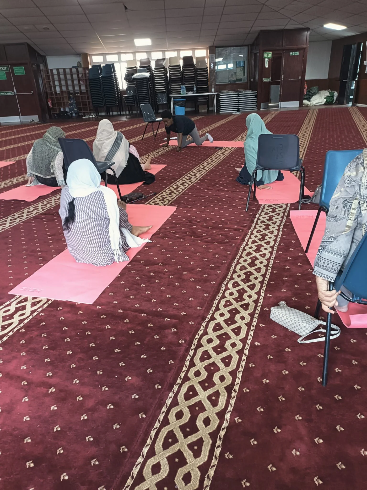 People practicing yoga on pink mats inside a spacious room with red patterned carpet, chairs, and stacked tables in the background.