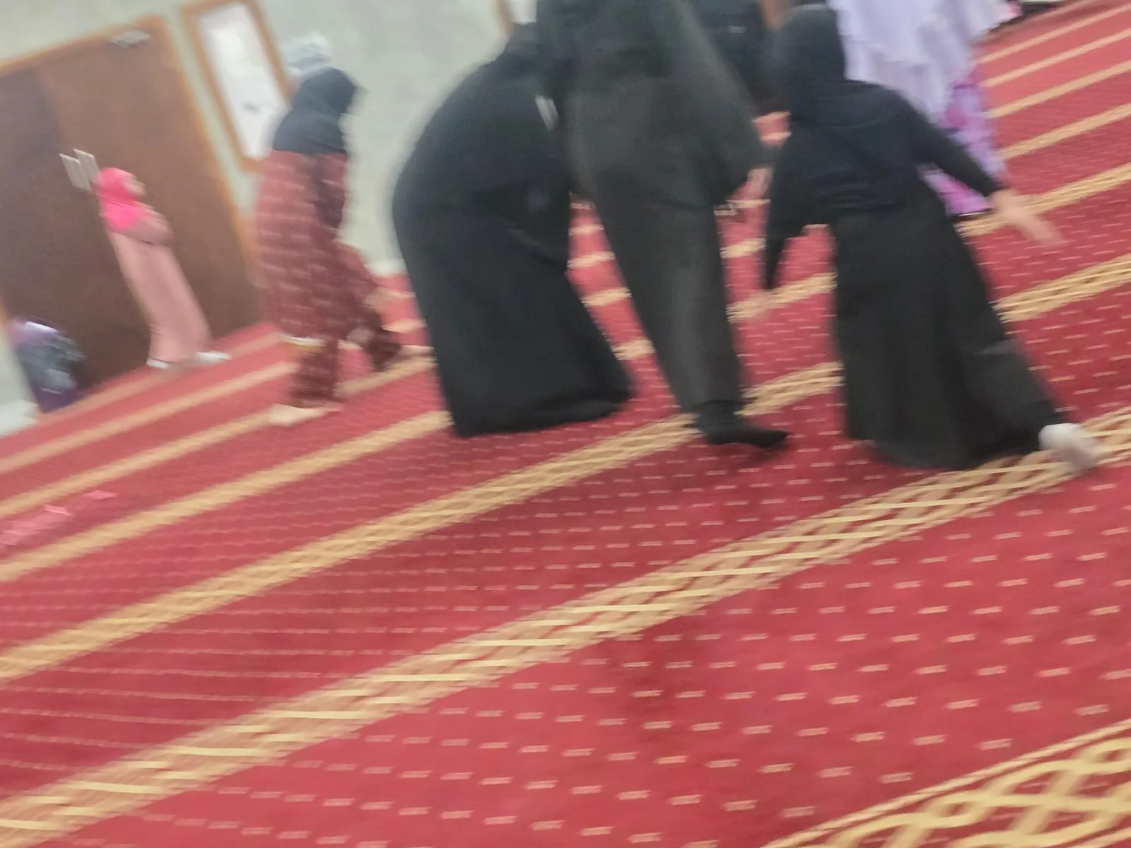 People kneeling and praying on a red and gold patterned carpet in a mosque.
