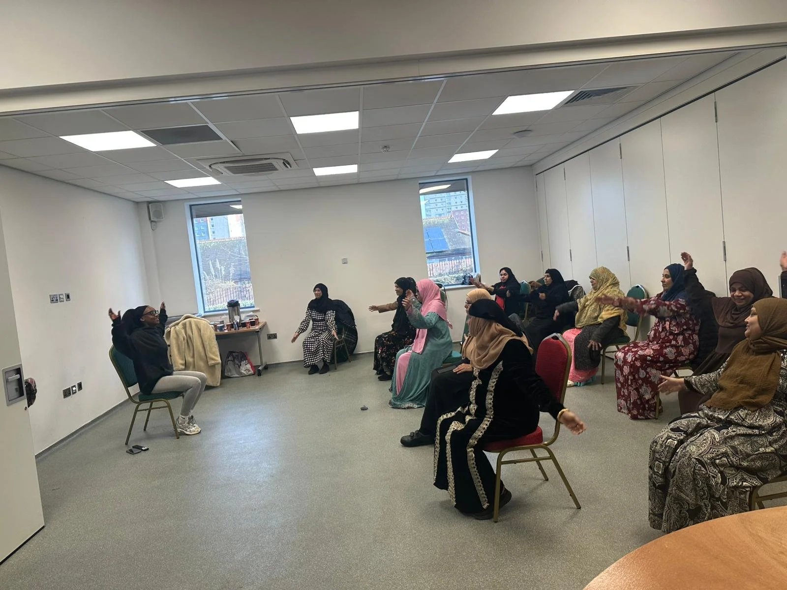 Women participating in a seated group activity in a spacious, well-lit conference room with two large windows showing a cityscape outside.