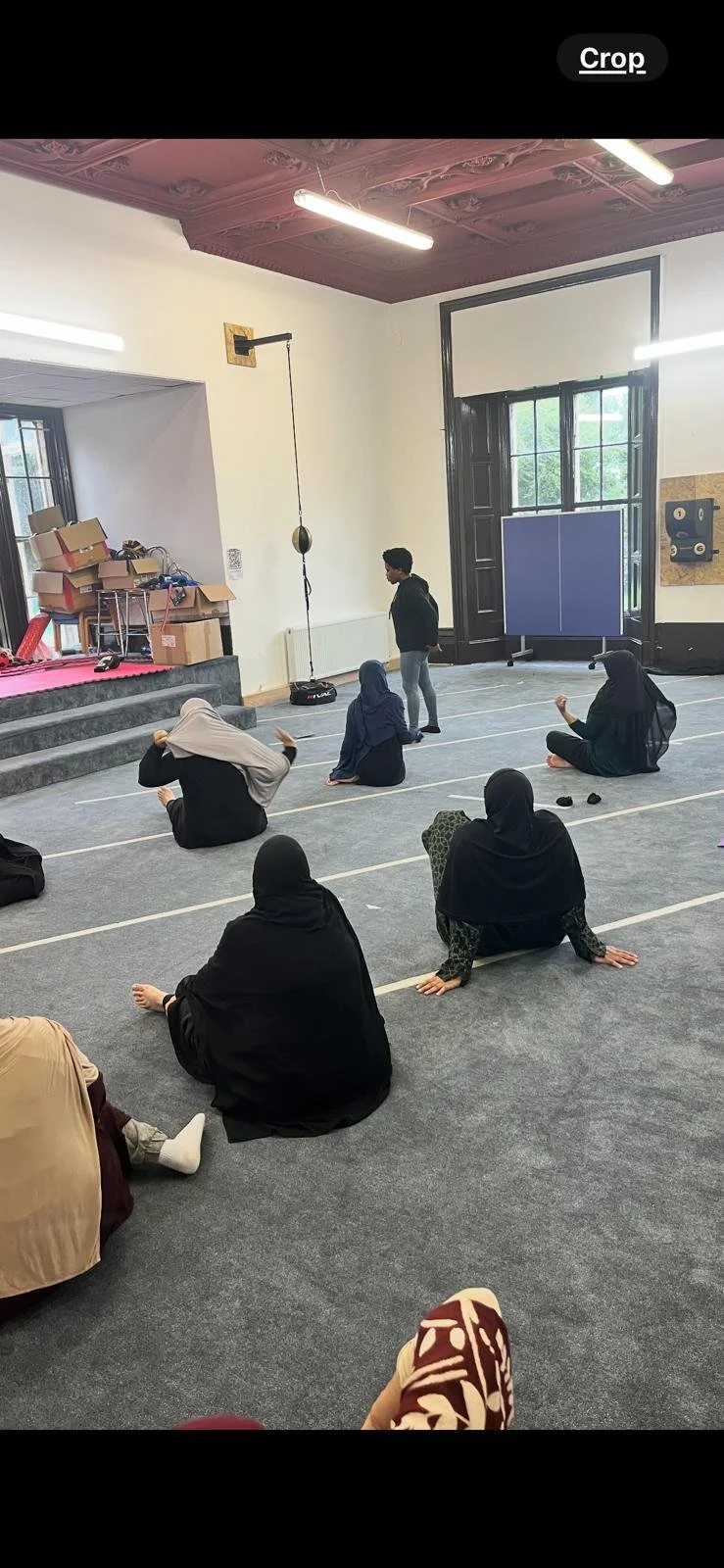 People participating in a religious or cultural class, sitting on the floor in a room with high ceilings, large windows, and hanging equipment, with some individuals wearing head coverings.