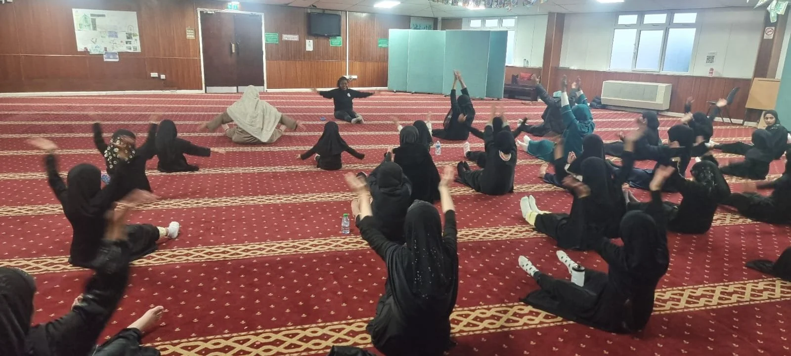 Group of children in black attire sitting on a red patterned carpet in a room, participating in a seated exercise or activity, with a woman leading the session at the front.