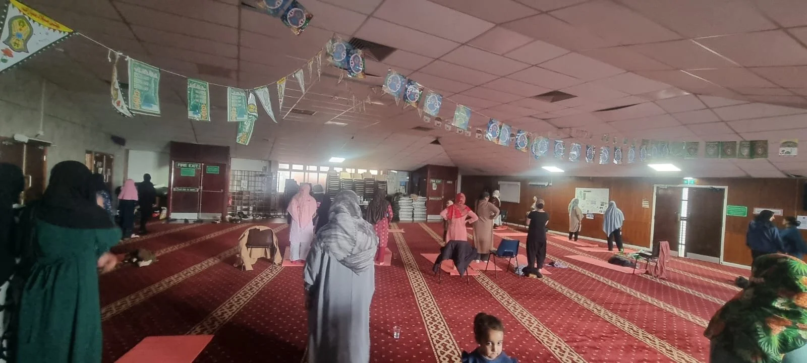 Women inside a mosque or prayer hall during prayer time, with some standing and some sitting on the red carpet, decorated with banners and flags hanging from the ceiling.