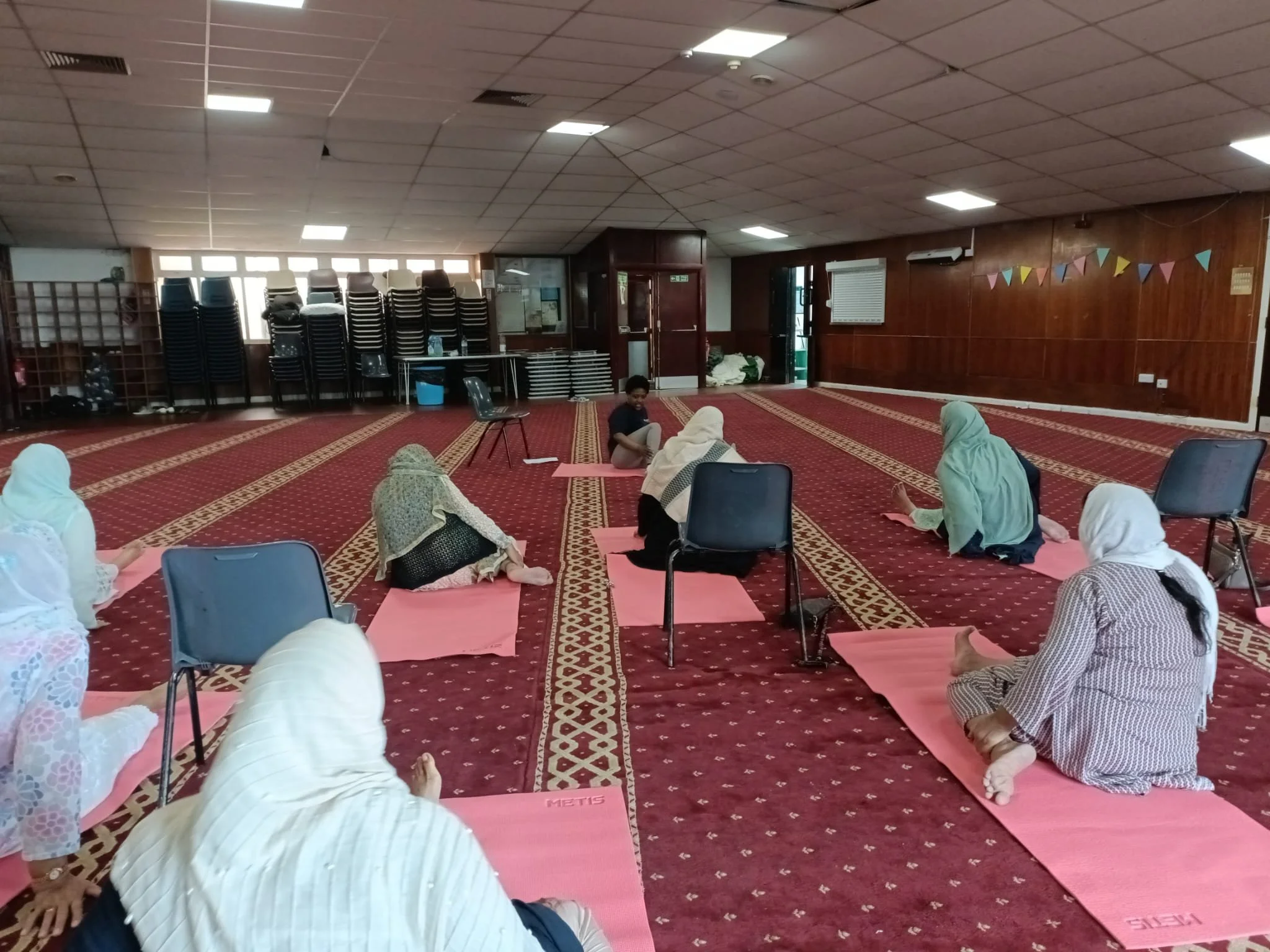 People participating in a seated yoga or meditation class on pink mats in a large indoor space with red carpet and wooden walls.