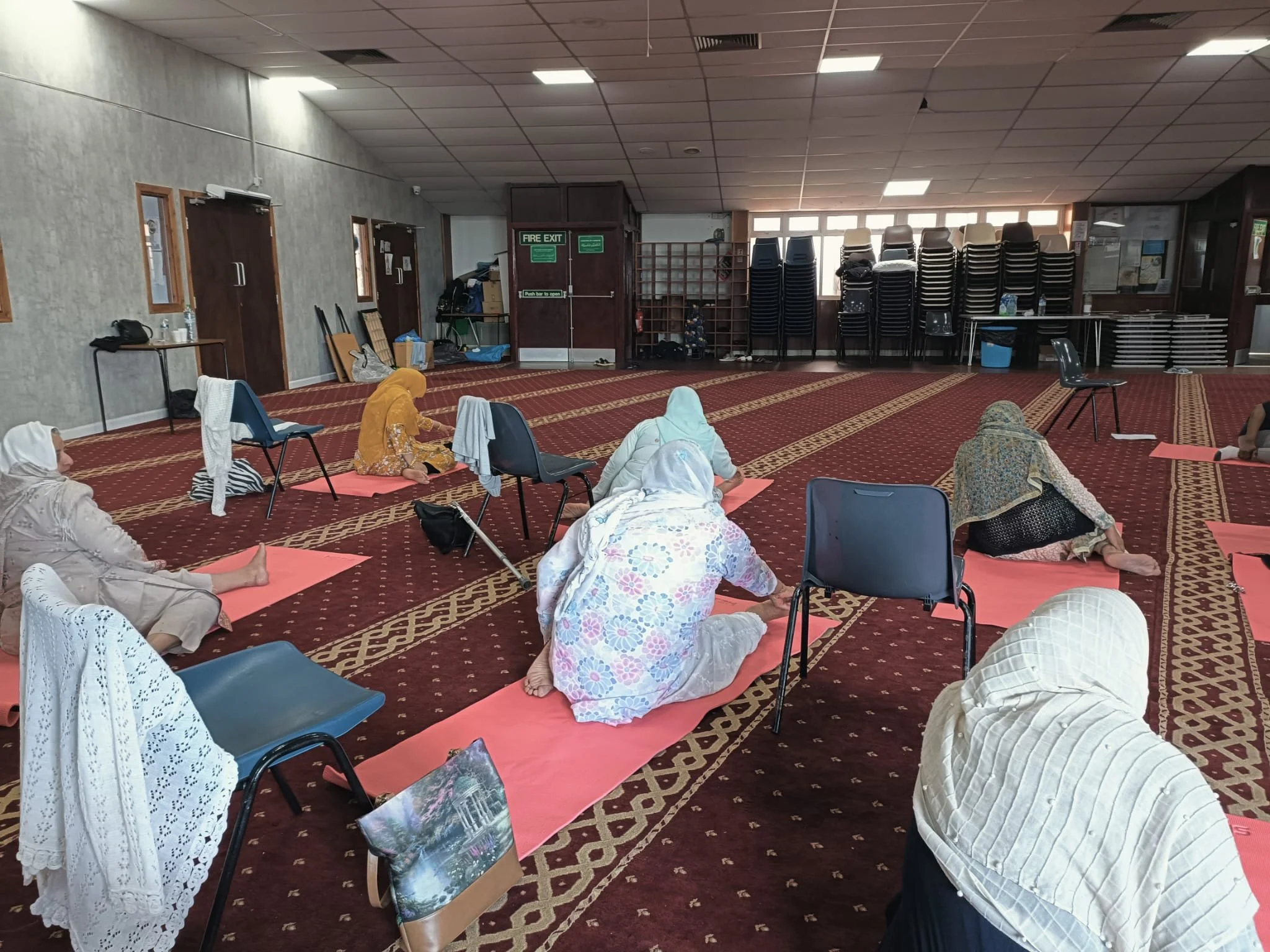 People participating in a seated yoga or meditation session on pink mats in a large room with chairs and stacked tables along the back wall.