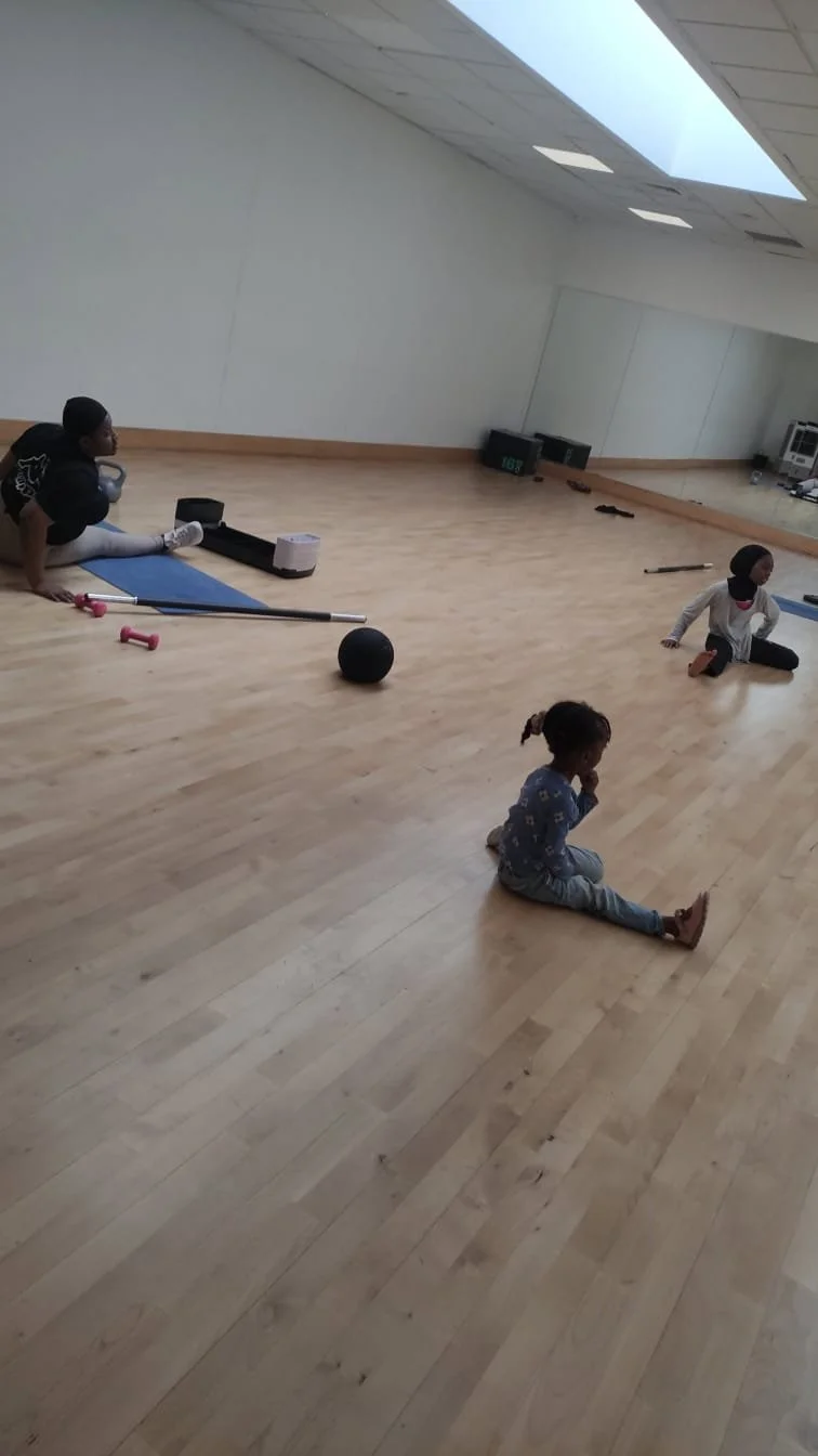 Children sitting on the wooden floor in a fitness or dance studio with high skylight, mirrors, and exercise equipment.