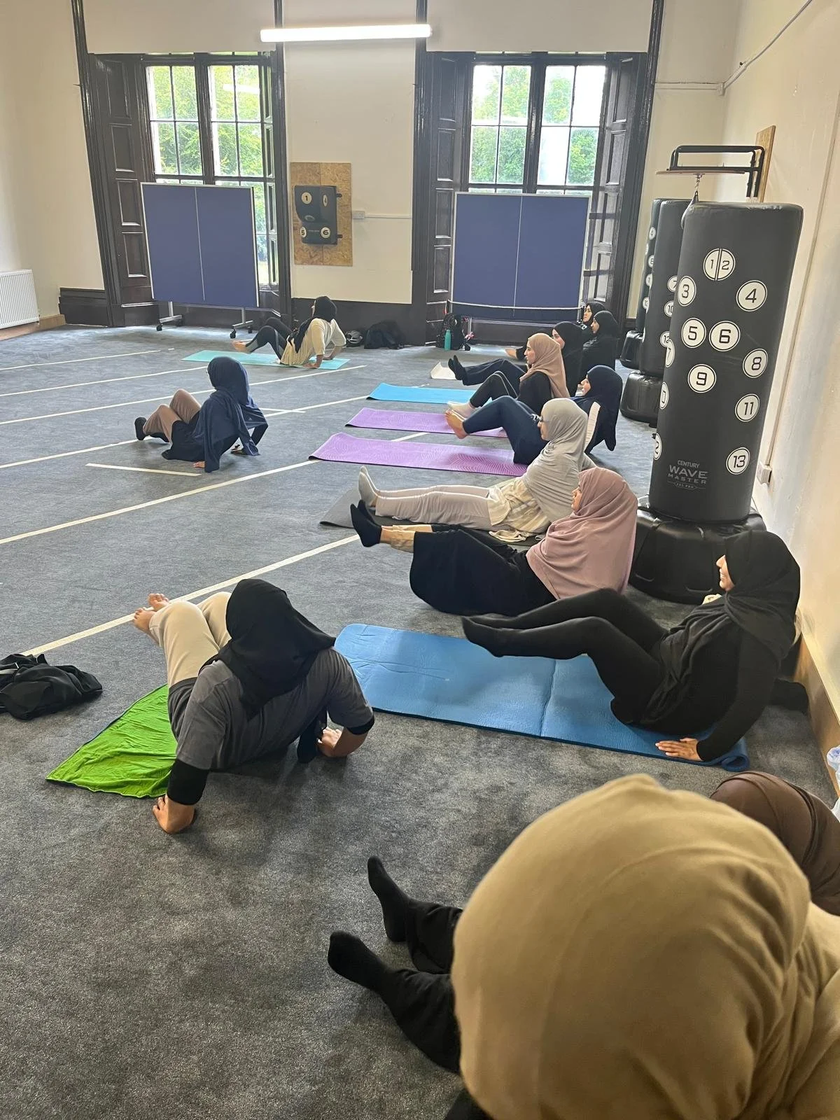 Women in hijabs participating in a fitness or yoga class in a spacious room with large windows, exercise mats, and fitness equipment.