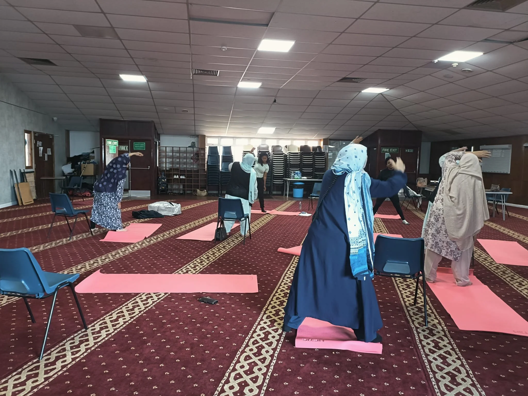 A group of women practicing yoga in a spacious indoor room with chairs and stacked tables in the background. They are on pink yoga mats, some with chairs for support, and several are wearing headscarves.