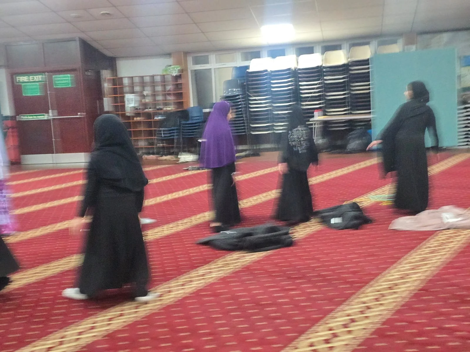 Women wearing black hijabs and clothing, standing and walking in a prayer area with red patterned carpet, in a mosque. Some have luggage or bags on the floor, and there are stacked chairs and shelves in the background.