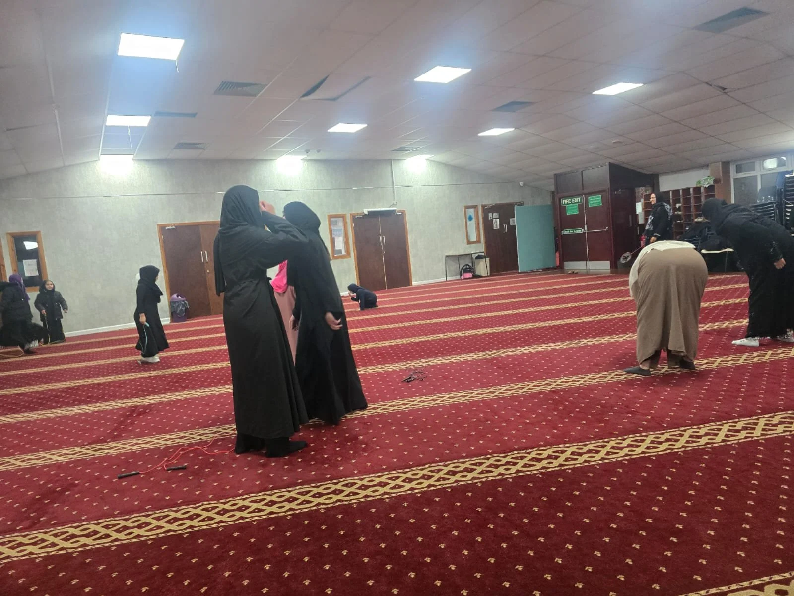 People praying inside a mosque, standing on a red patterned carpet with gold accents, some bowed and others in supplication, with a gray wall and doors in the background.