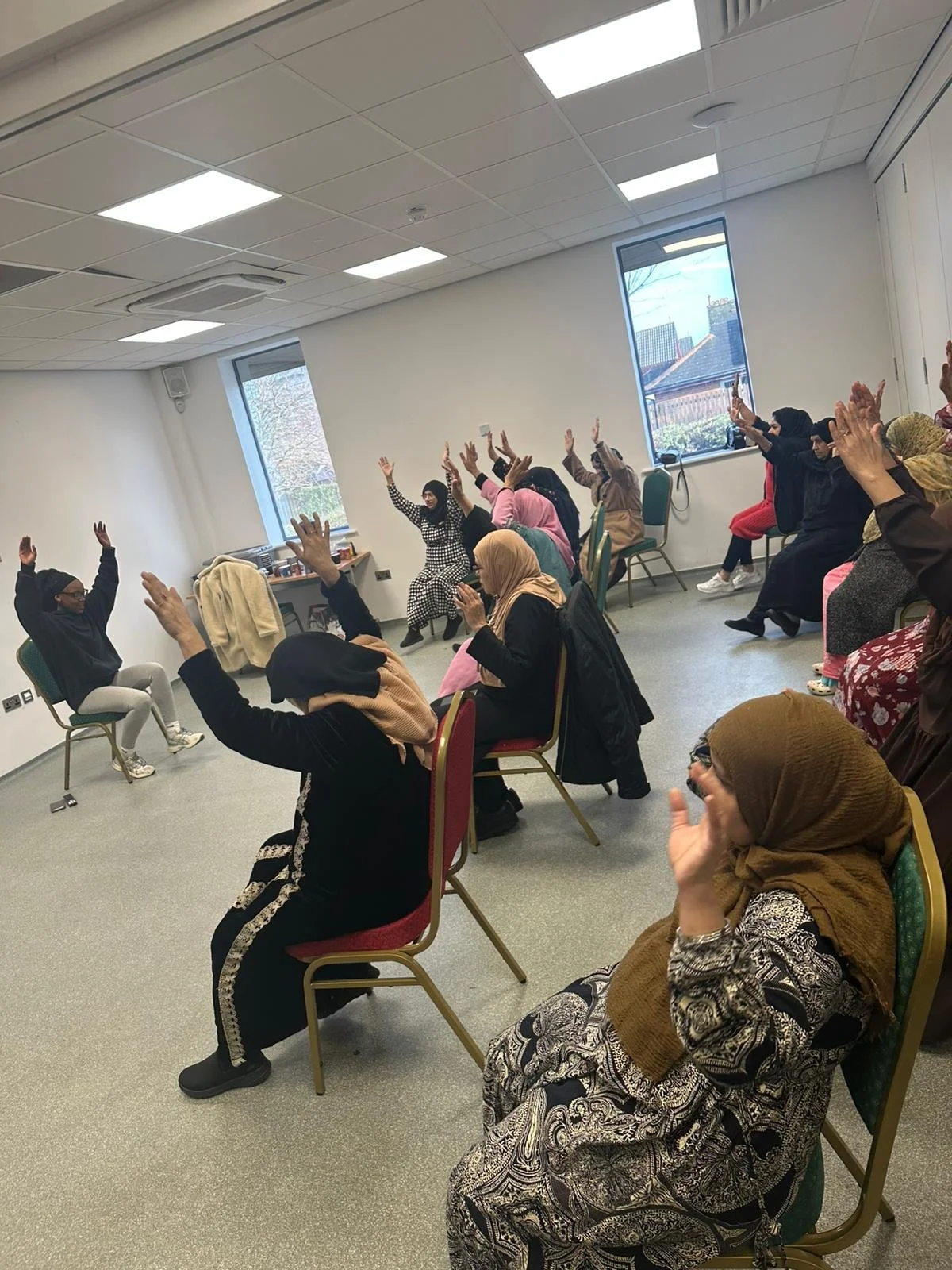 Women seated in a room with large windows raising their hands, participating in an activity or class.