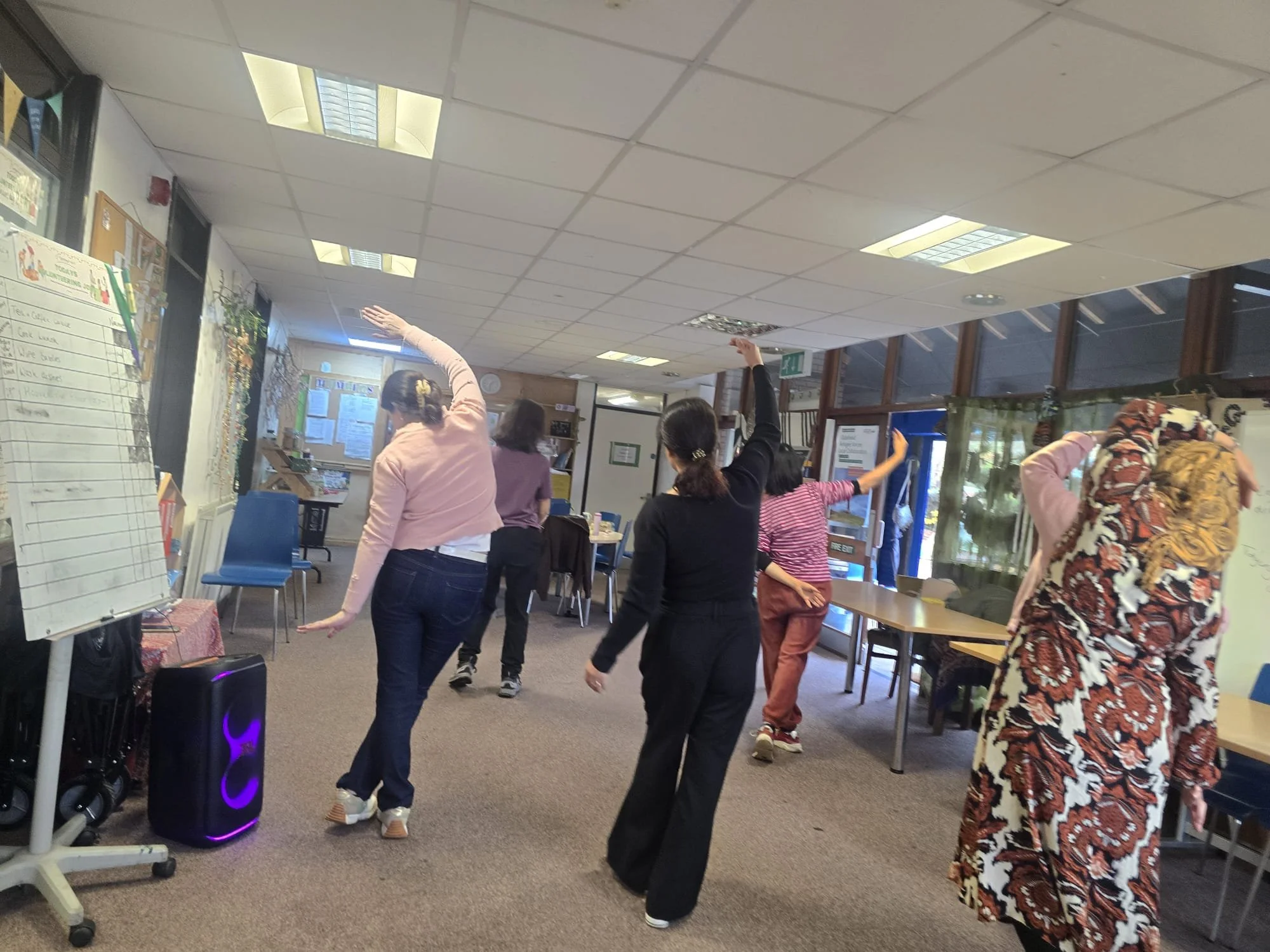 Group of women practicing dance moves in an indoor community room, with tables, chairs, and a whiteboard in the background.