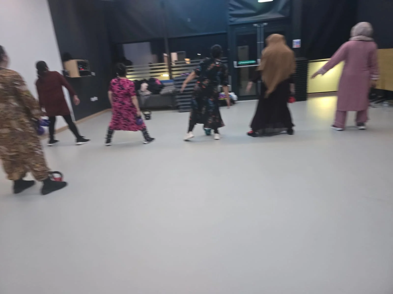 Group of people walking indoors in a line, some wearing colorful dresses, in a spacious room with dark walls and a gray floor.