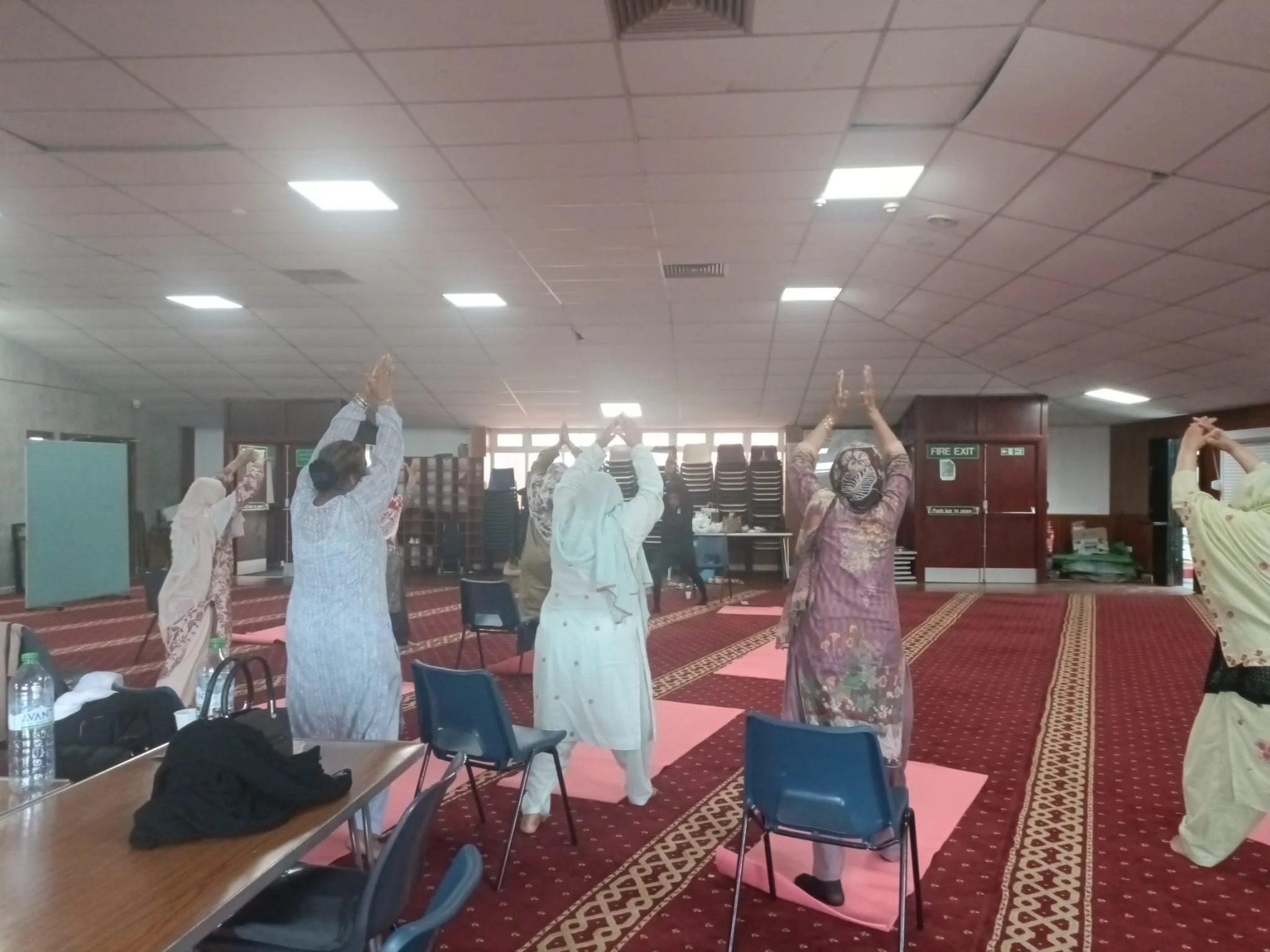 A group of women wearing traditional dress participating in a yoga or stretching class in a large, carpeted room with chairs, tables, and stacked chairs in the background.