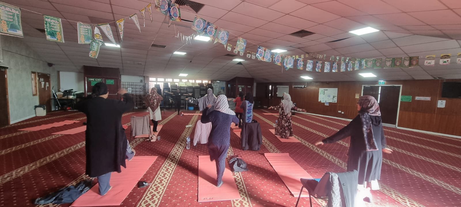 Women in a spacious mosque practicing yoga on pink mats, with prayer rugs and banners hanging from the ceiling.