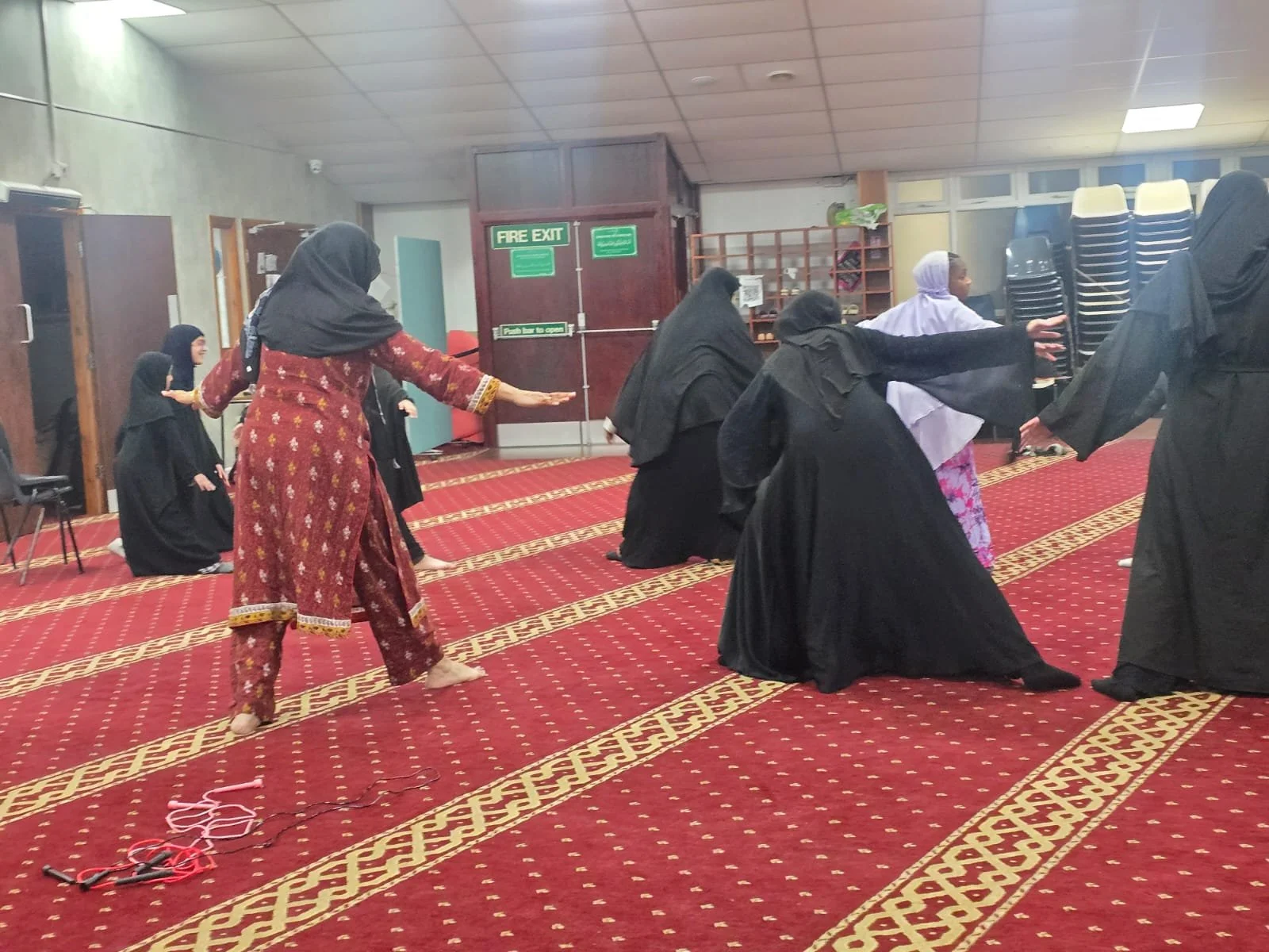 A group of women in traditional Muslim attire practicing prayer inside a mosque, with some women kneeling and others standing with outstretched hands on red patterned carpet.