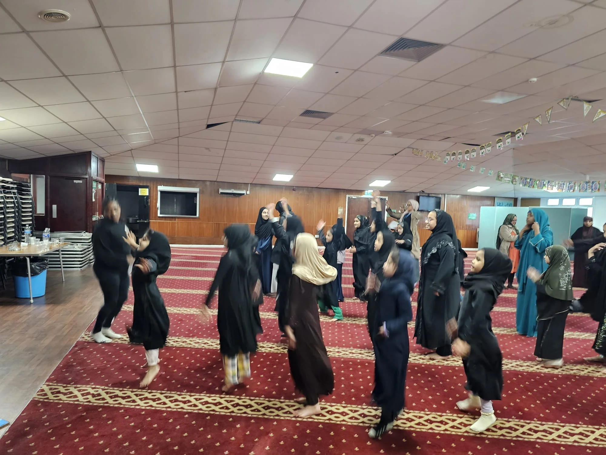Group of women dancing and enjoying themselves indoors, with some wearing traditional hijabs and modest clothing, in a hall with wooden wall panels and a red carpeted floor.