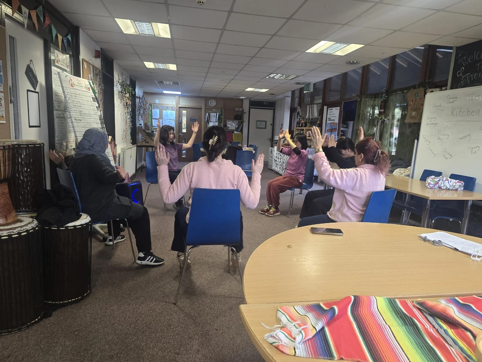 A group of women in a circle participating in a seated activity or dance in a room, with some raising their arms. There is a whiteboard with notes on the right side of the room and tables with colorful cloths.