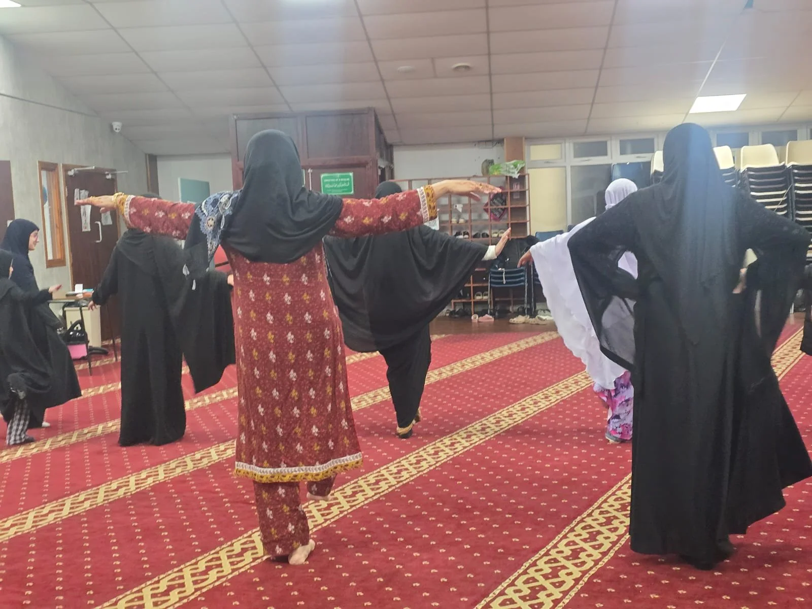 Women in traditional Islamic dress performing a dance or exercise in a mosque with a red carpet and stacked chairs in the background.