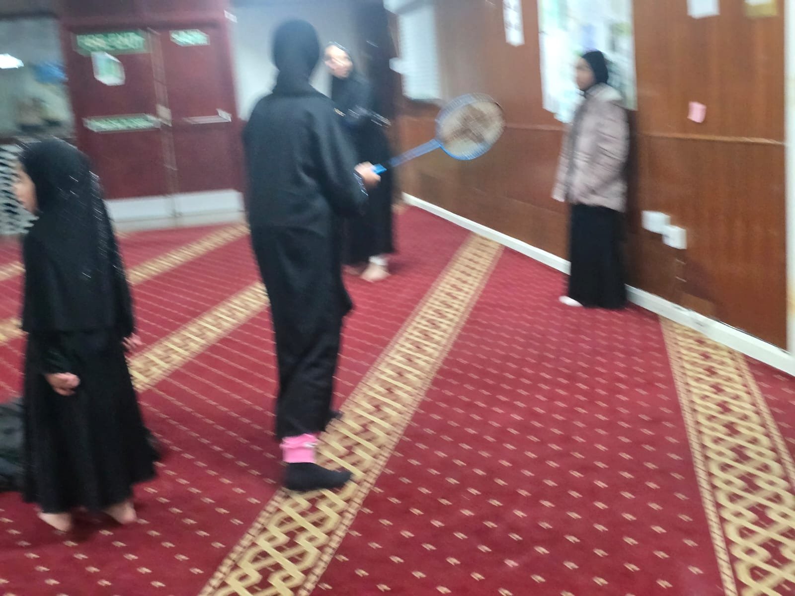 People in a room, some wearing black robes and hijabs, with one person holding a badminton racket and a woman standing against a wooden-paneled wall.