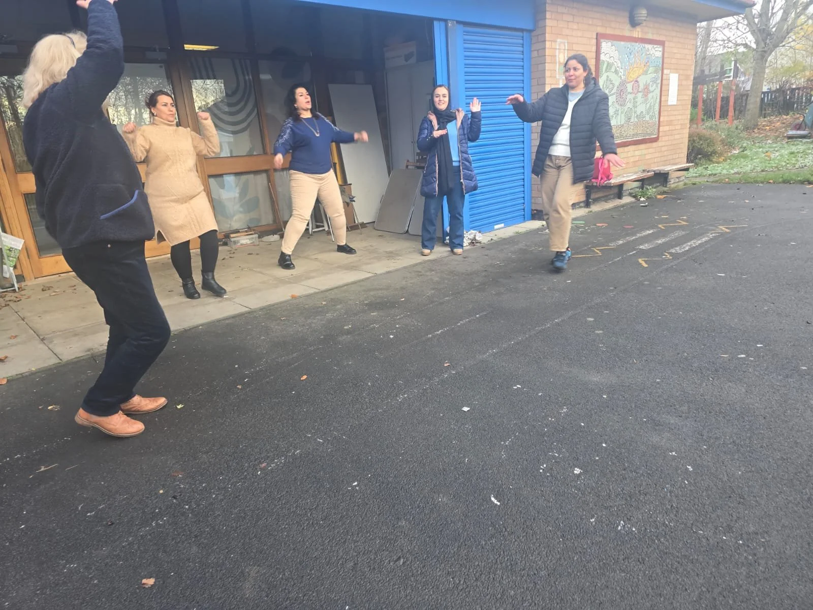 Five women are outside, dancing and having fun near a building with a colorful mural. Some are wearing jackets, and the ground is paved.