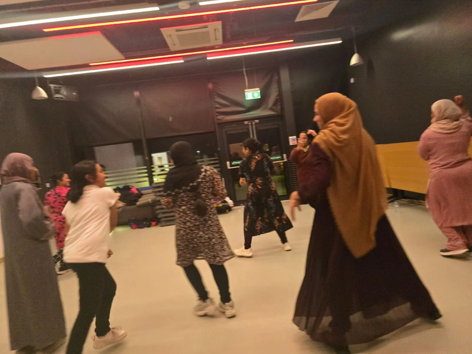 Group of women of diverse backgrounds and wearing headscarves dancing and enjoying themselves in an indoor space with a dark wall and large windows.