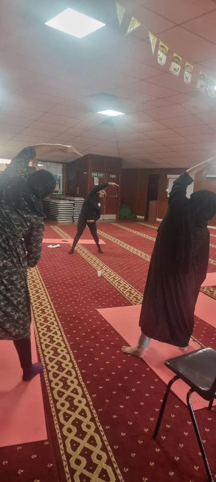 Women practicing yoga in a mosque, standing on pink mats, with prayer rugs and flags hanging from the ceiling.