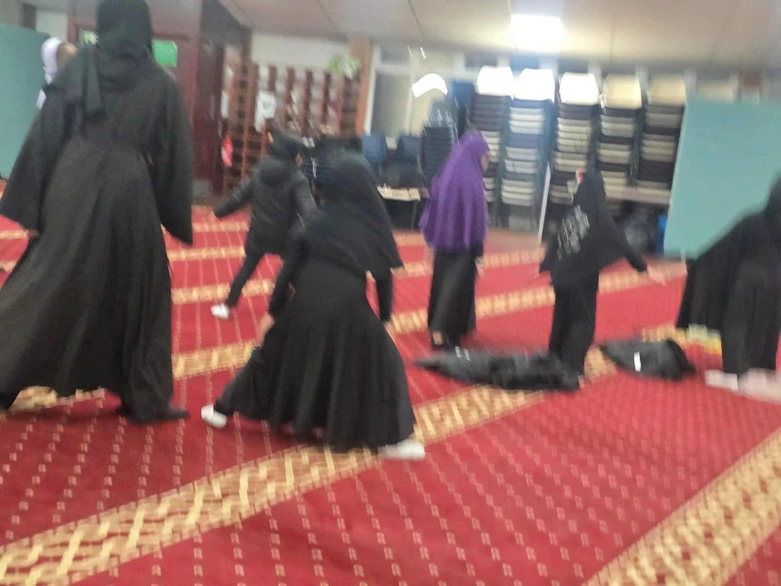 Several mannequins dressed in black in a room with red and gold patterned carpet, with stacked chairs and a green sign in the background.