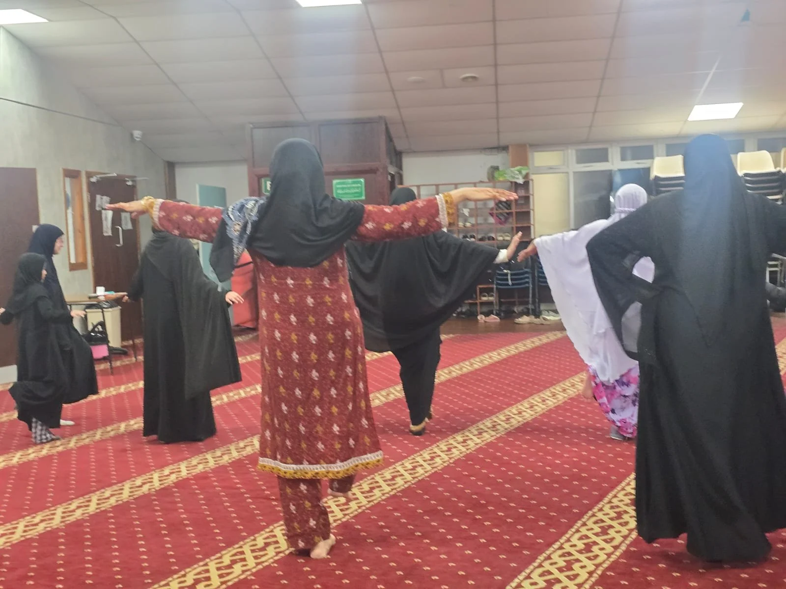 Women in traditional Islamic attire participating in a communal prayer or dance in a hall with a red patterned carpet, some with arms extended.