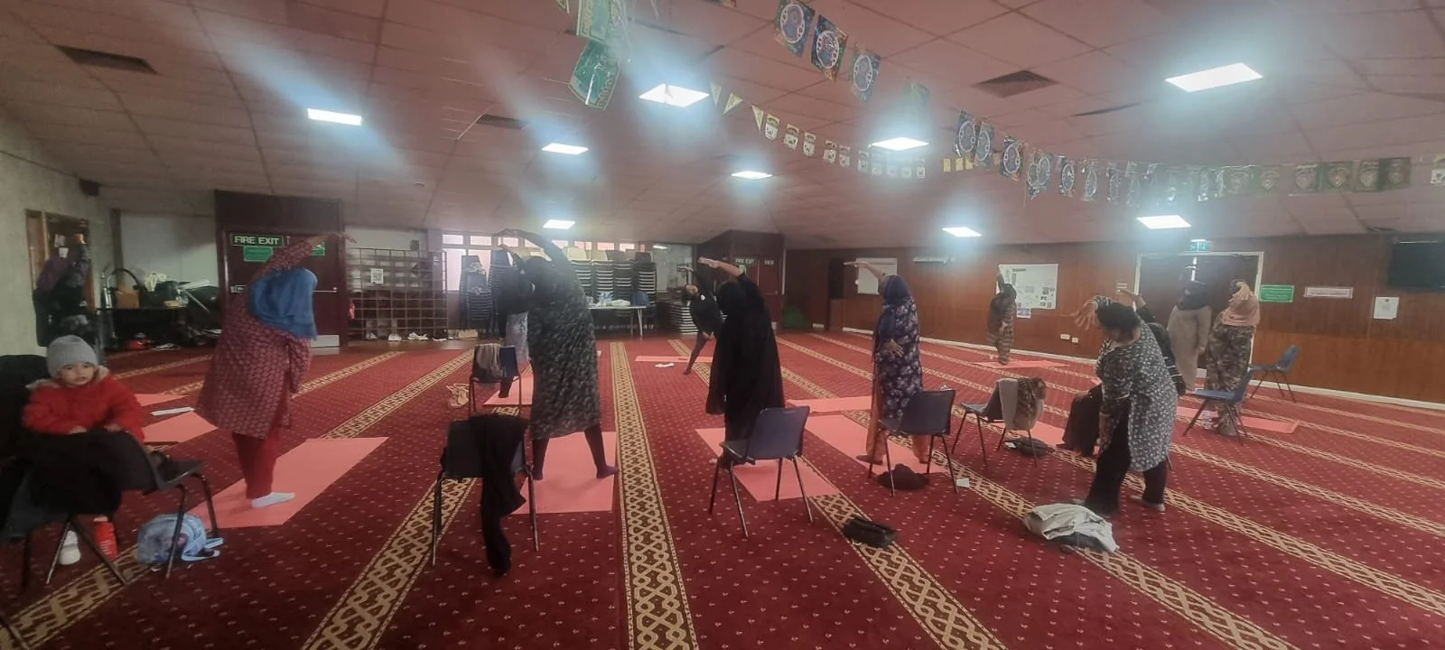 People practicing social distancing and prayer in a mosque with chairs and prayer mats on a red patterned carpet, hanging decorations and a fire exit sign in the background.