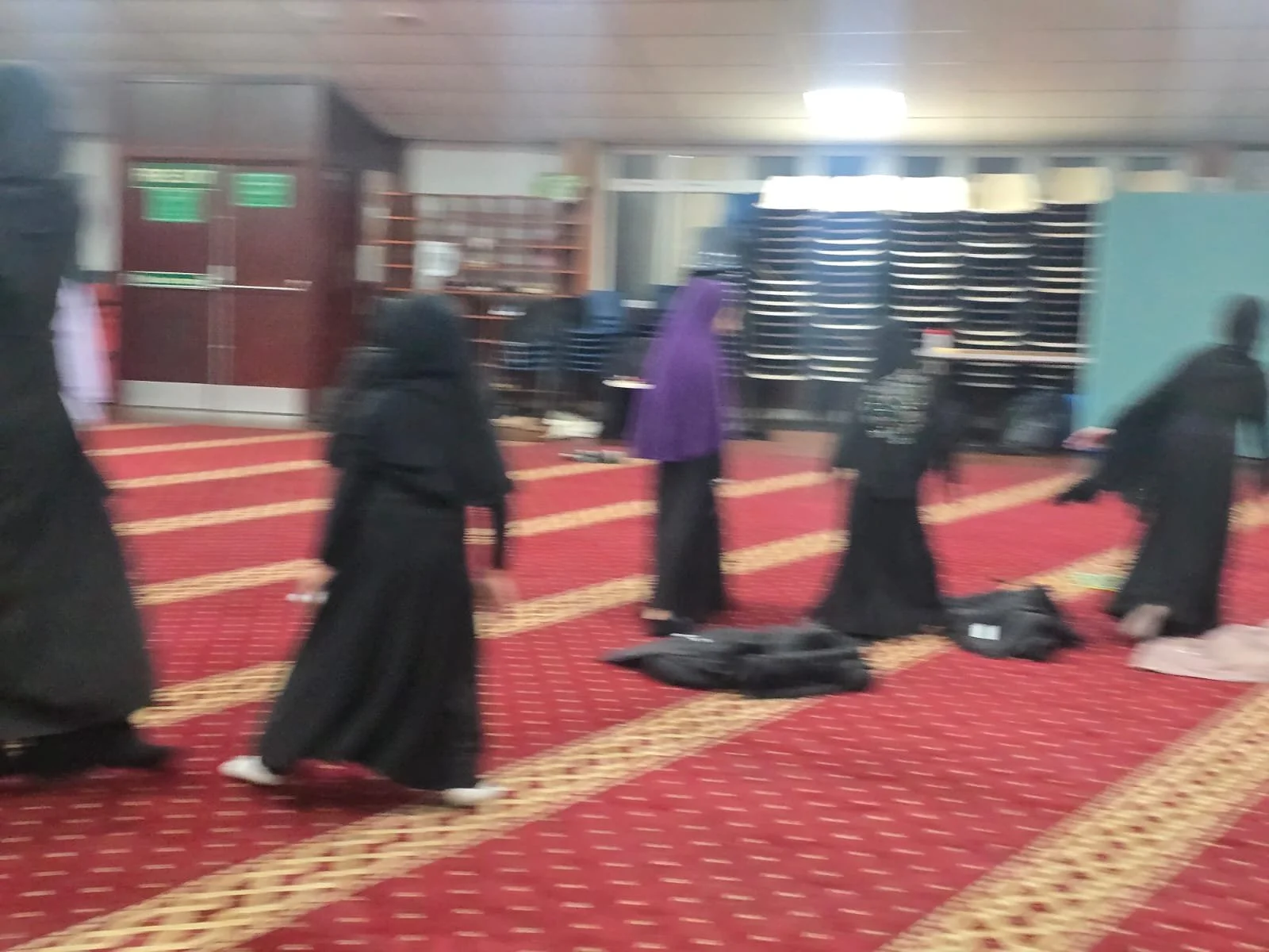 People in prayer inside a mosque, some standing and some kneeling on prayer rugs.