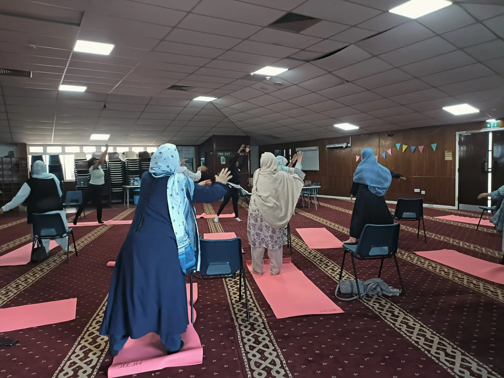 Women practicing yoga in a community center with pink yoga mats, chairs, and bunting on the wall.