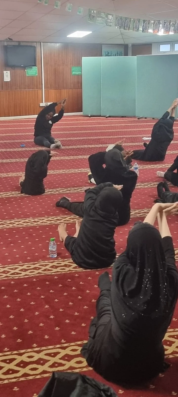 A group of women wearing black hijabs and modest clothing sitting on a red patterned carpet, participating in a yoga or stretching session inside a room with wood-paneled walls and hanging banners.