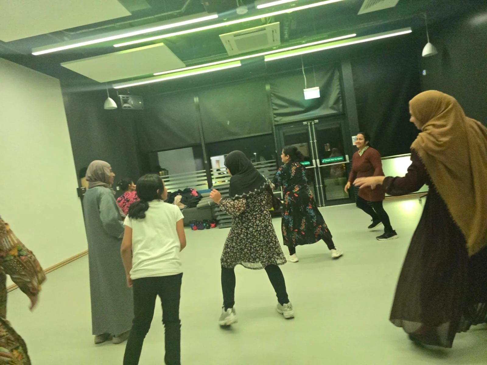 Women and girls dancing in a spacious indoor room with dark walls and ceiling, illuminated by green and white lights.