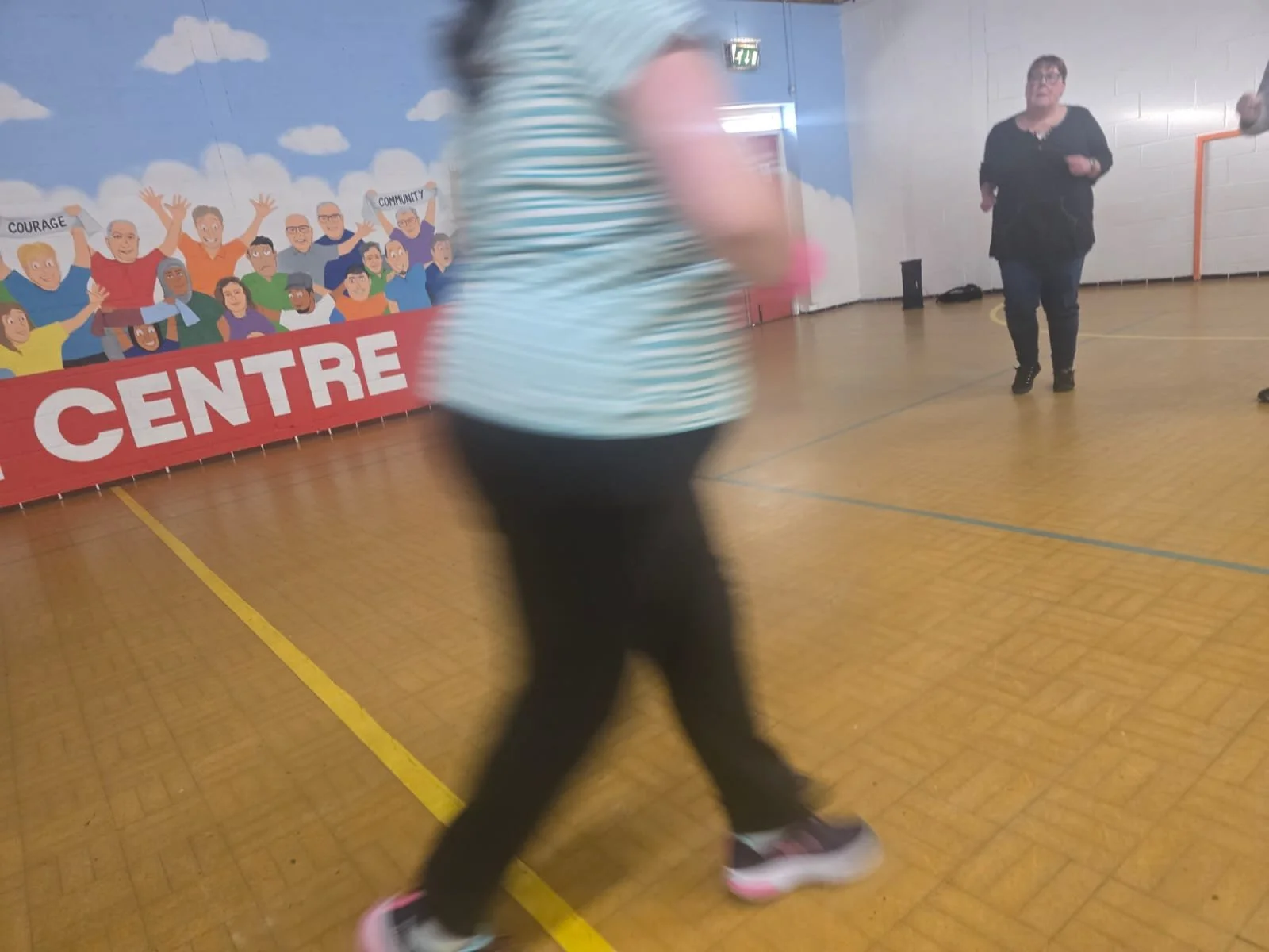 A blurred person in a striped shirt walking inside a gymnasium with a mural of diverse animated characters holding signs that read words like 'Courage' and 'Community'.