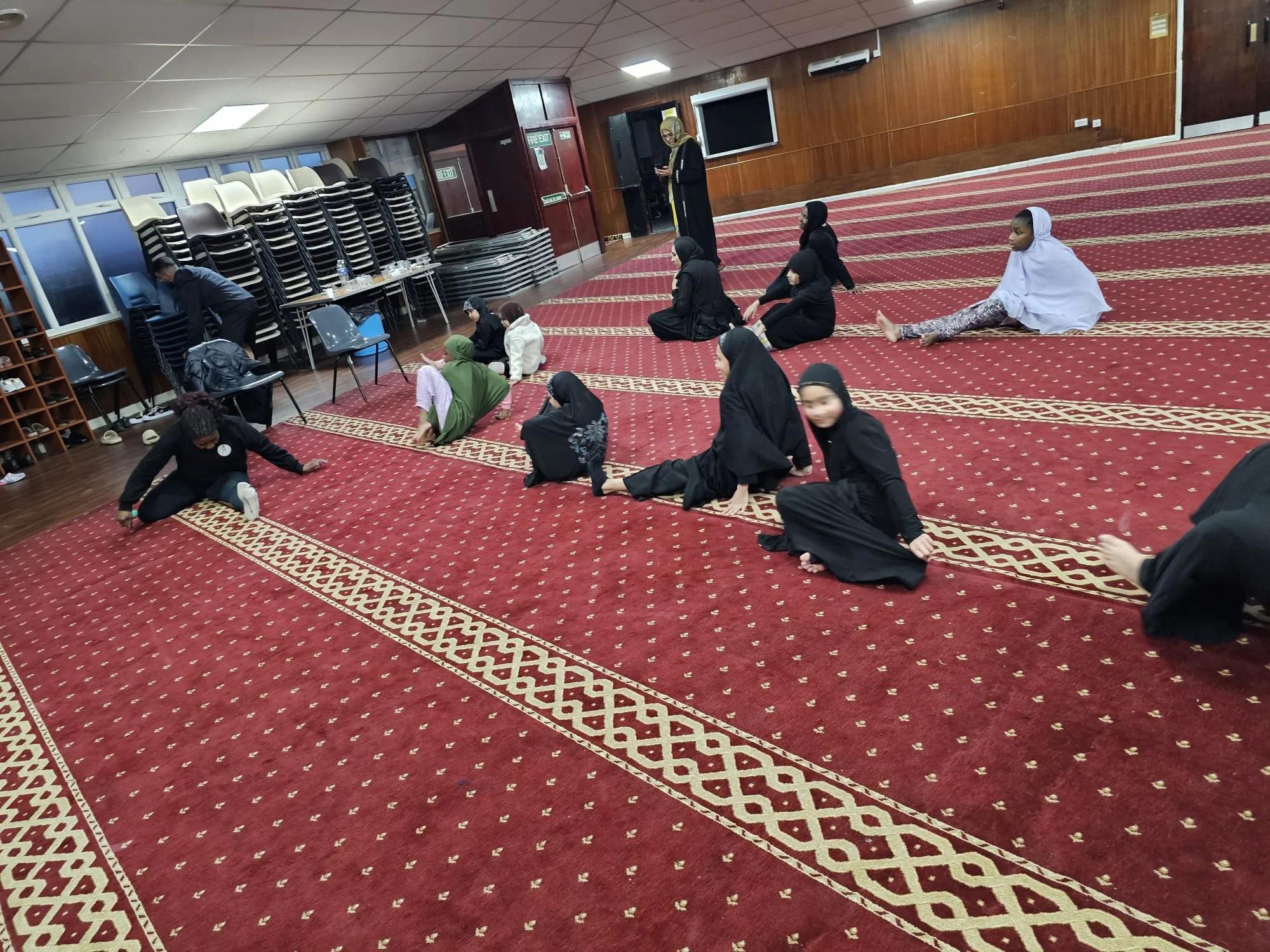Group of women and children practicing a seated exercise in a mosque prayer hall with red carpet and wooden walls.