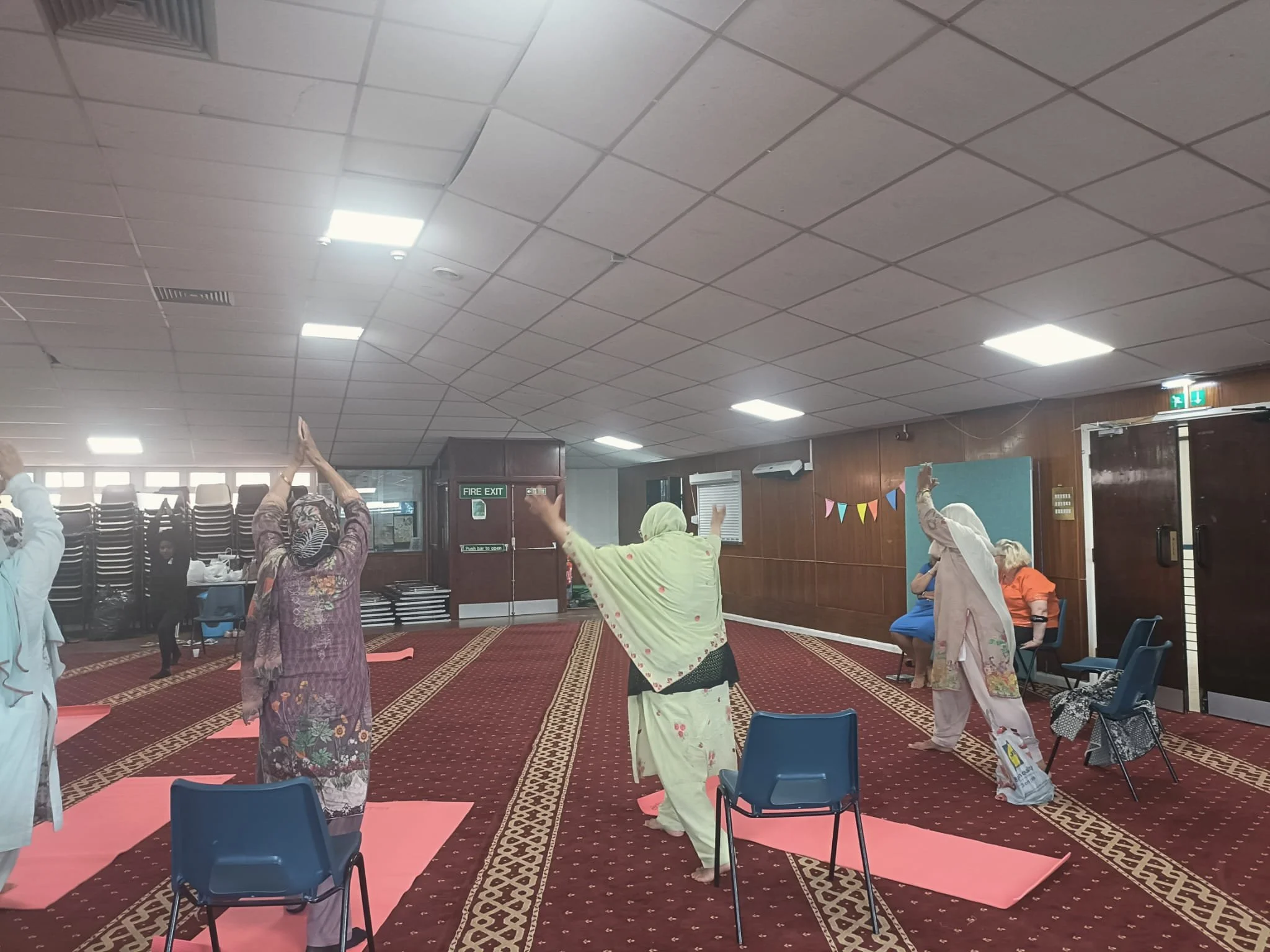 Group of people practicing yoga on pink mats in a community room with chairs on the side, decorated with colorful bunting and a fire exit door.