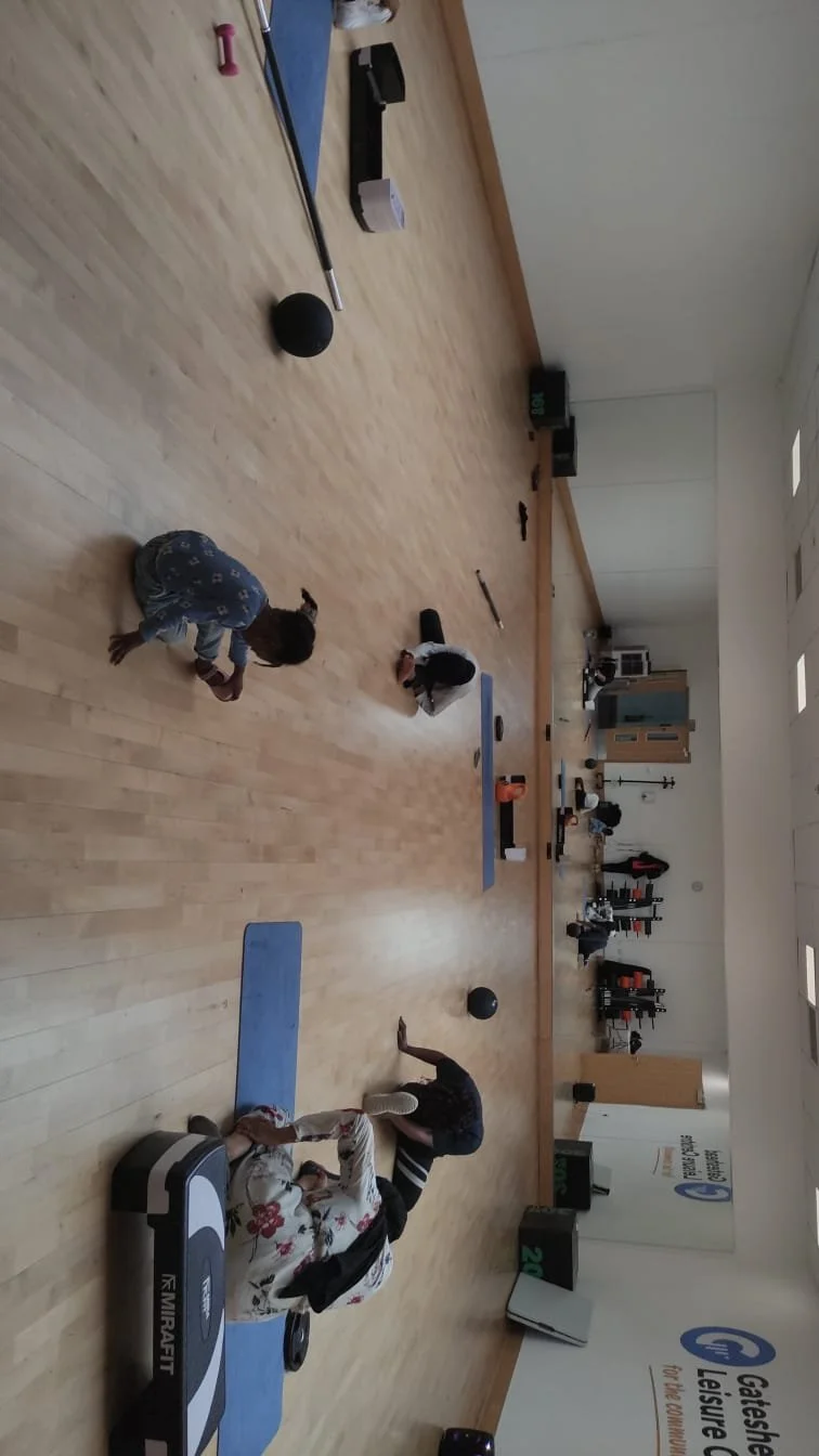 Two children in a gym or activity room, one crawling on the floor and the other kneeling, with various exercise equipment scattered around, including a yoga mat, medicine ball, and foam roller.