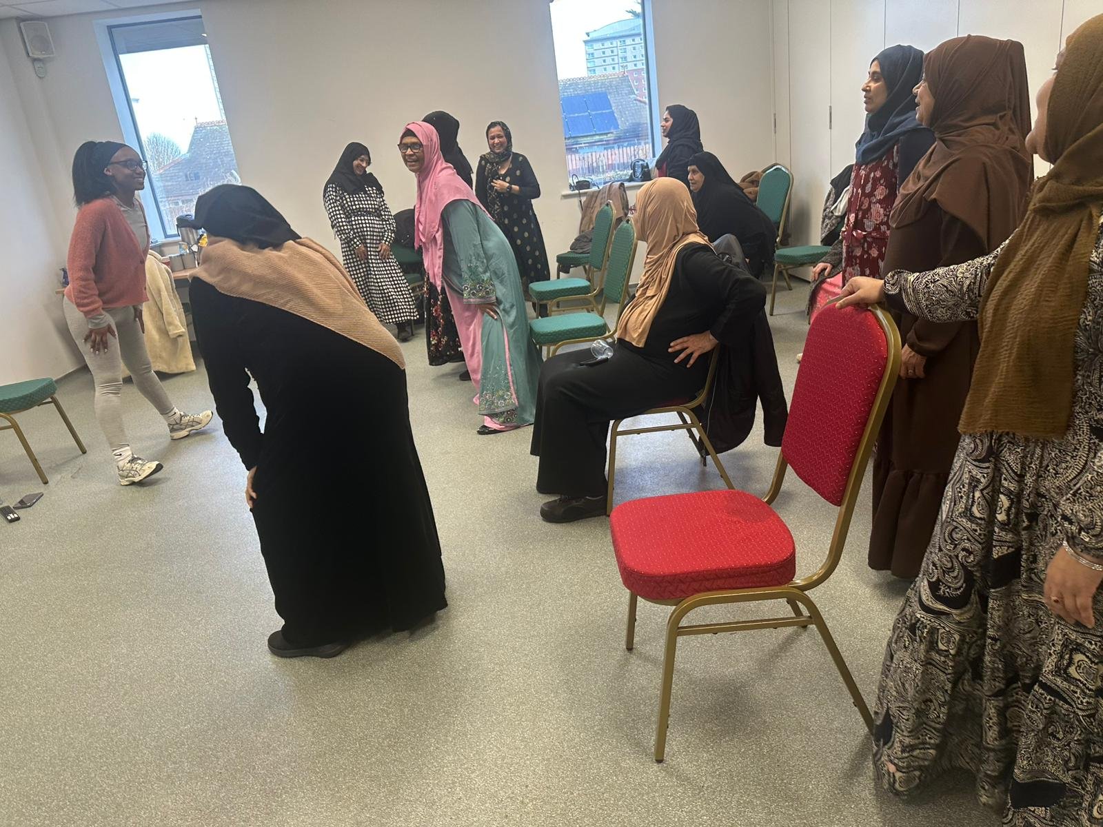 Group of women in a room, some standing and some sitting, participating in a activity or workshop. Several women are wearing traditional hijabs and long dresses, with a few in casual clothing. The room has a window showing buildings outside and is eq