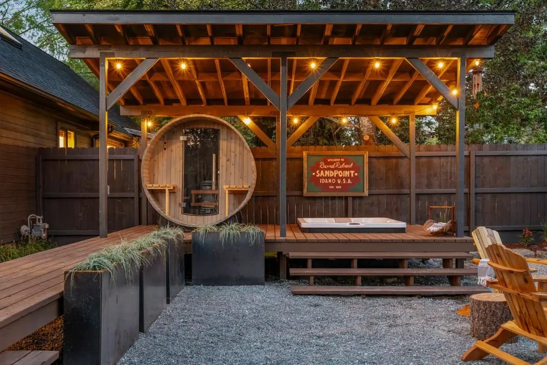 Outdoor backyard space with a wooden pavilion featuring string lights, a round hot tub, Adirondack chairs, and a sign that says "Welcome to Sandpoint, Idaho U.S.A." with gravel ground and trees in the background.