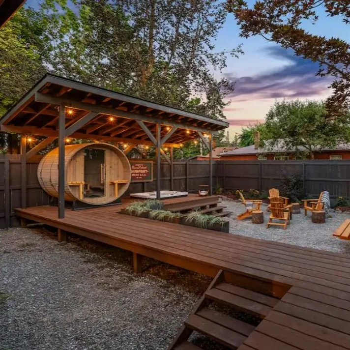 Backyard space with wooden deck, a sauna barrel, outdoor seating with wooden chairs, and string lights, surrounded by a wood fence and trees at sunset.