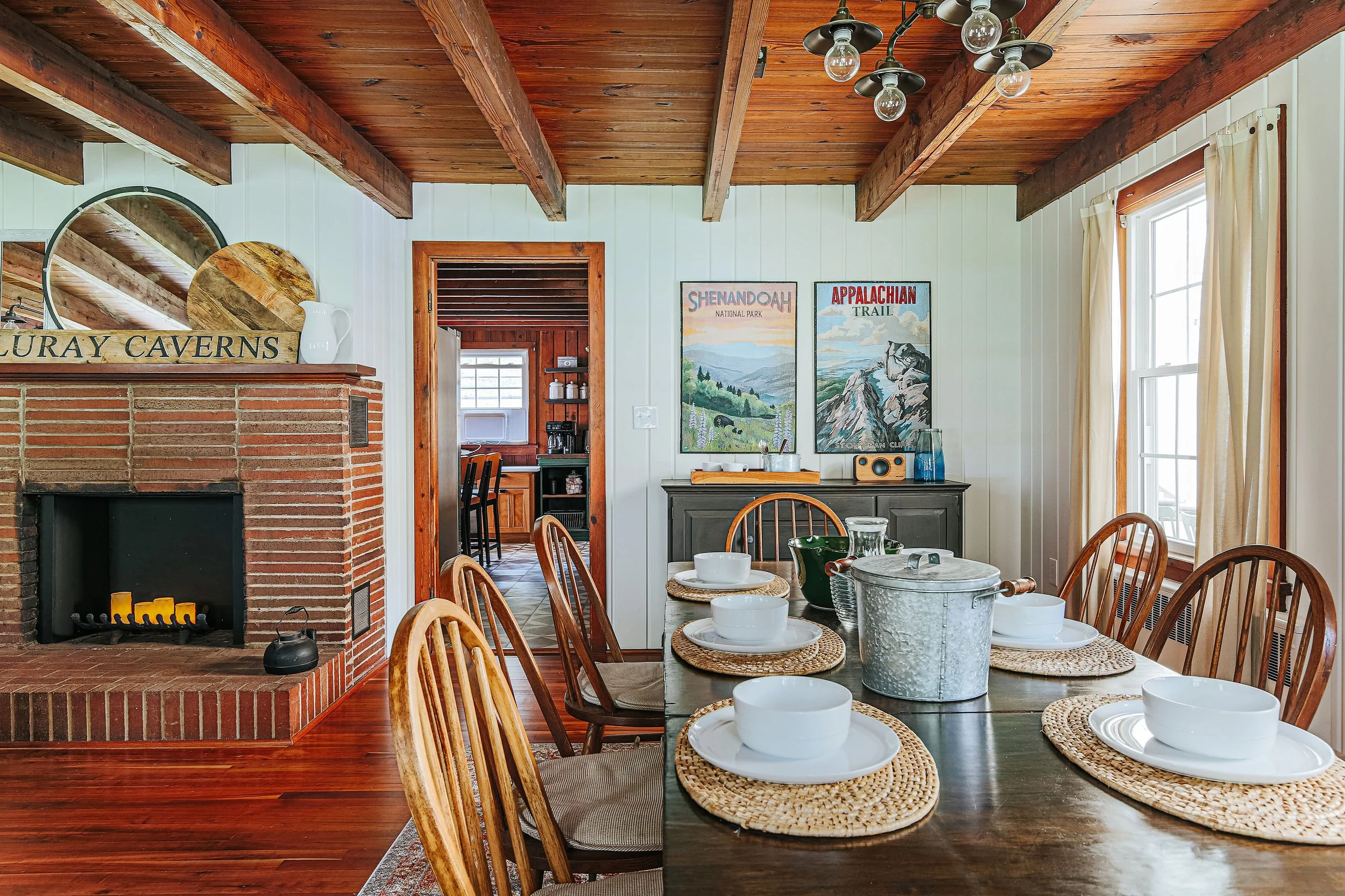 A cozy dining room with a wooden table set with white bowls, plates, and woven placemats, surrounded by wooden chairs. There is a brick fireplace on the left with mirrors above it, and two framed landscape posters on the wall. The room has white paneled walls, a window with cream curtains, and a ceiling with exposed wooden beams and a modern light fixture.