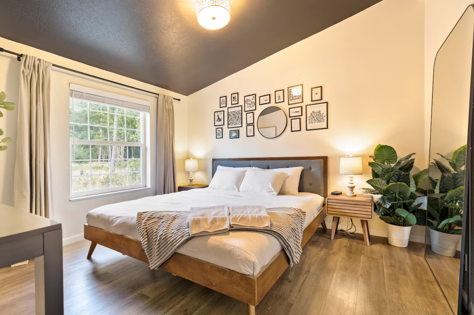 A cozy bedroom with a large bed, white bedding, and a blanket at the foot, flanked by two nightstands with lamps. The wall above the bed features a gallery of black-framed artwork and a round mirror. A large window with beige curtains reveals green trees outside. Two potted plants are in the corner. The ceiling is dark, contrasting with white walls, and the flooring is light wood.