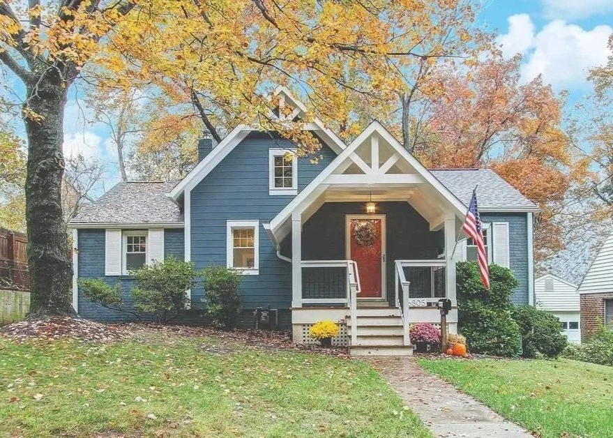 A blue house with white trim and a small front porch decorated with a wreath, autumn flowers, and a pumpkin. An American flag hangs beside the porch. The house is surrounded by bushes and trees with fall foliage.