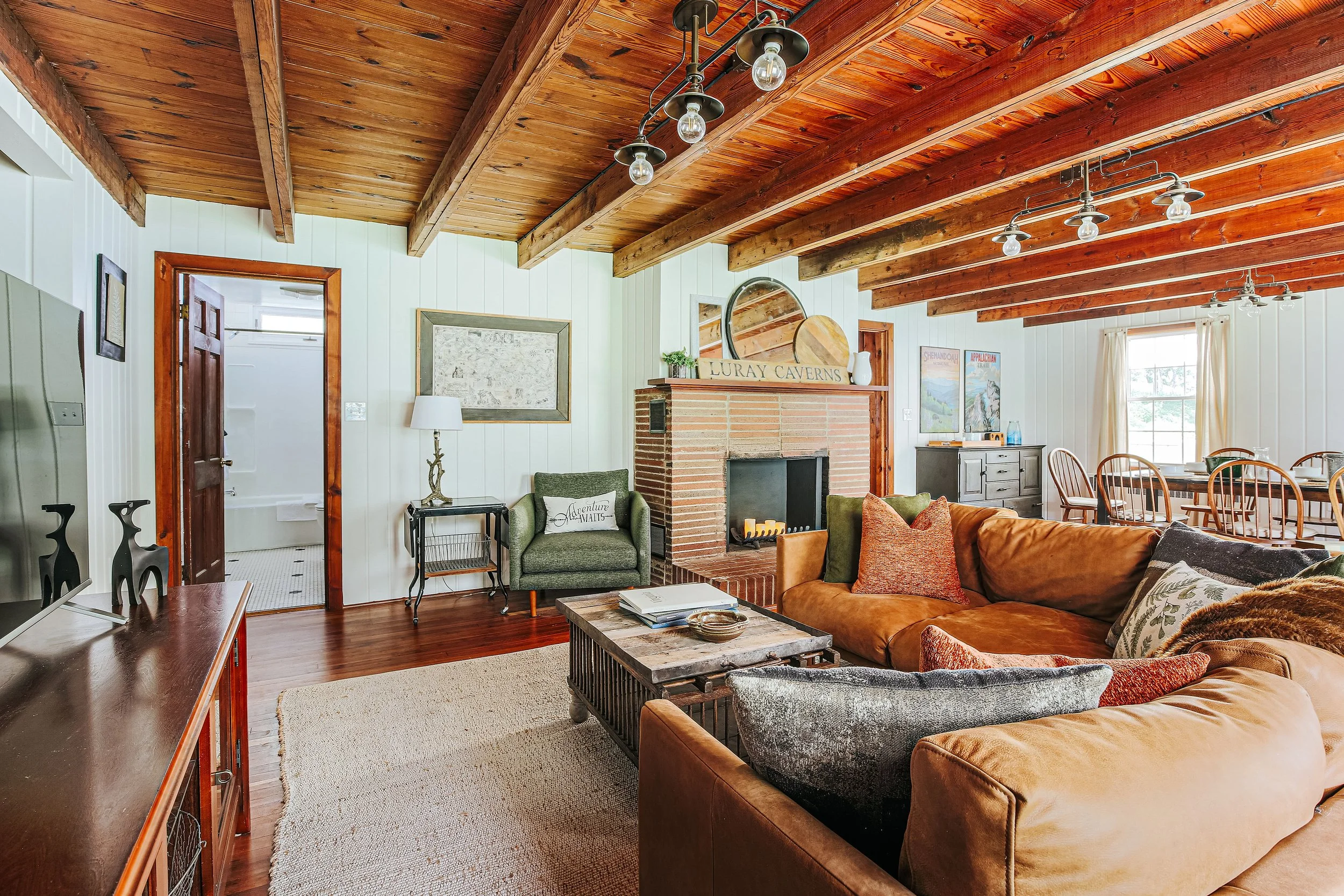 Living room with wooden ceiling beams, a brick fireplace, a beige couch, a green armchair, a coffee table, a TV, and a dining area with chairs and windows with curtains.