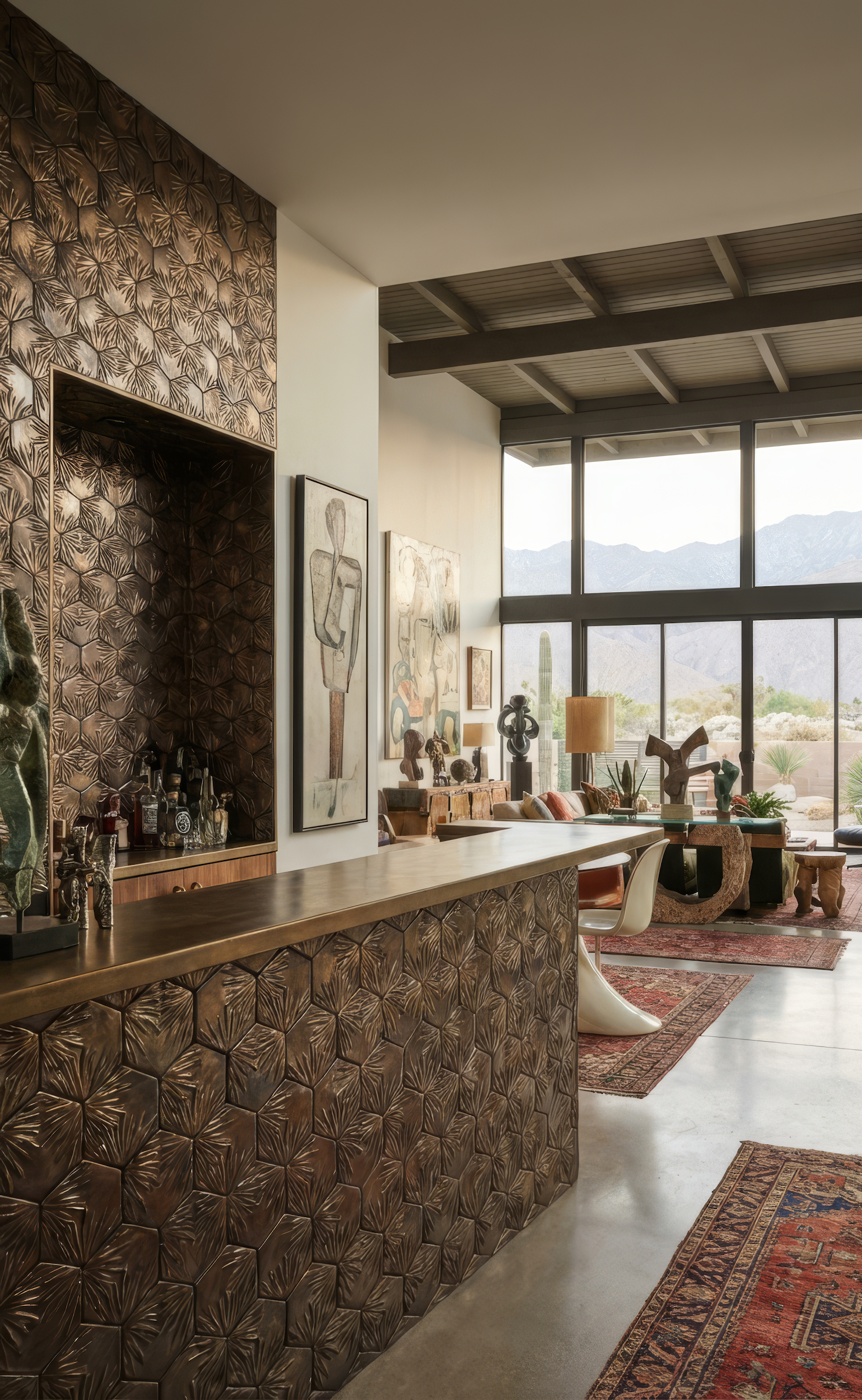 Interior of a modern living space with large windows showing desert mountains outside. Features include a textured bronze bar, eclectic artwork, a vintage-style rug, and various sculptures and plants.
