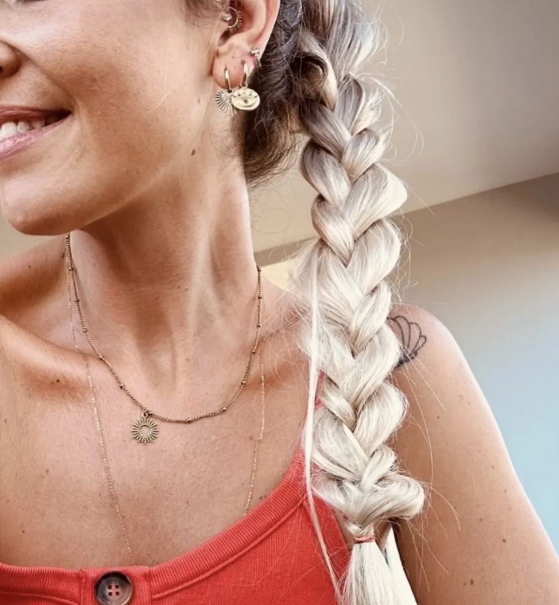 Partie d'une femme avec des cheveux blonds en tresse, portant des boucles d'oreilles, des colliers, et un haut rouge avec un bouton.